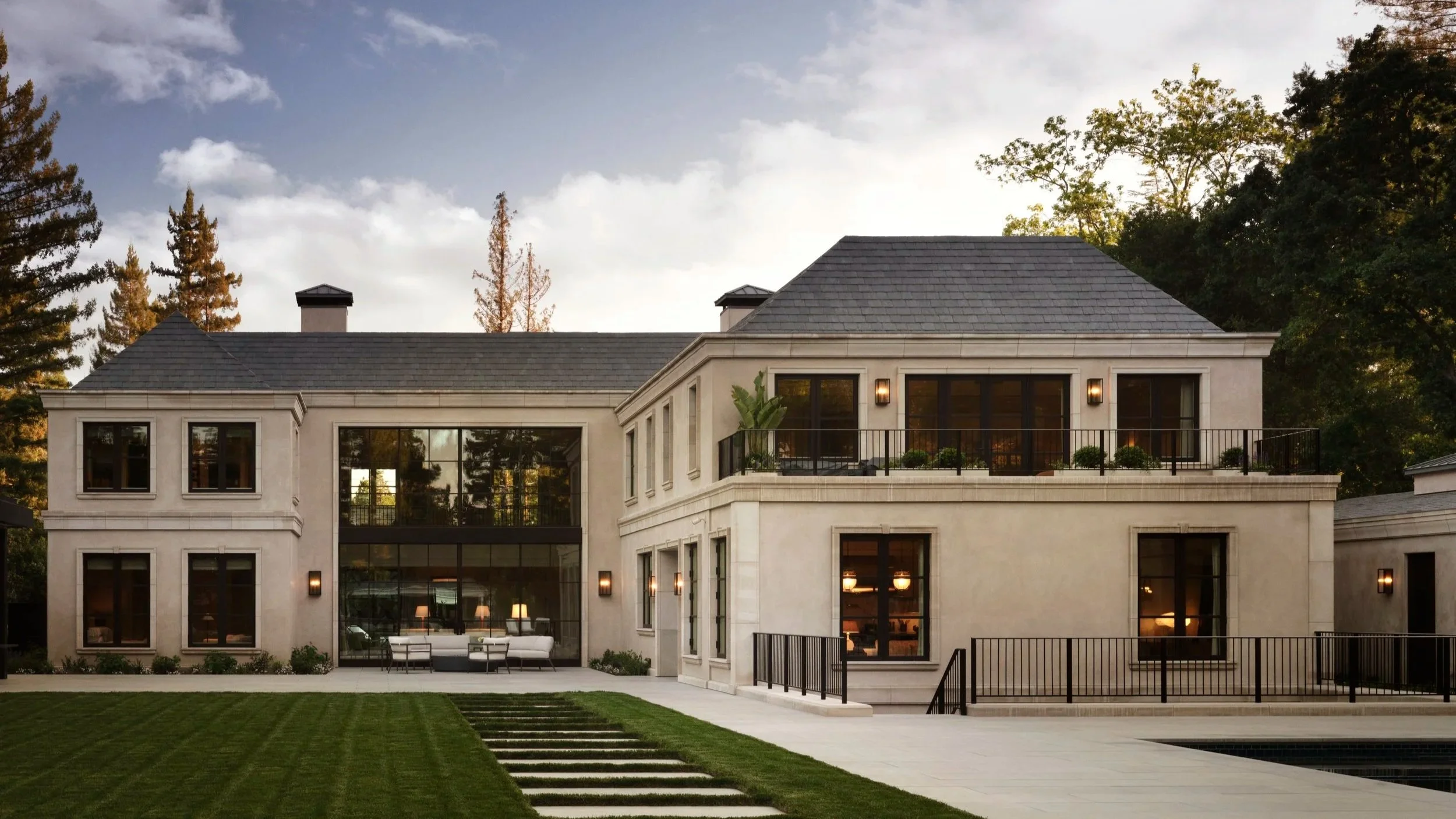 A modern two-story house with large windows, a landscaped lawn, and a patio with seating, surrounded by trees.