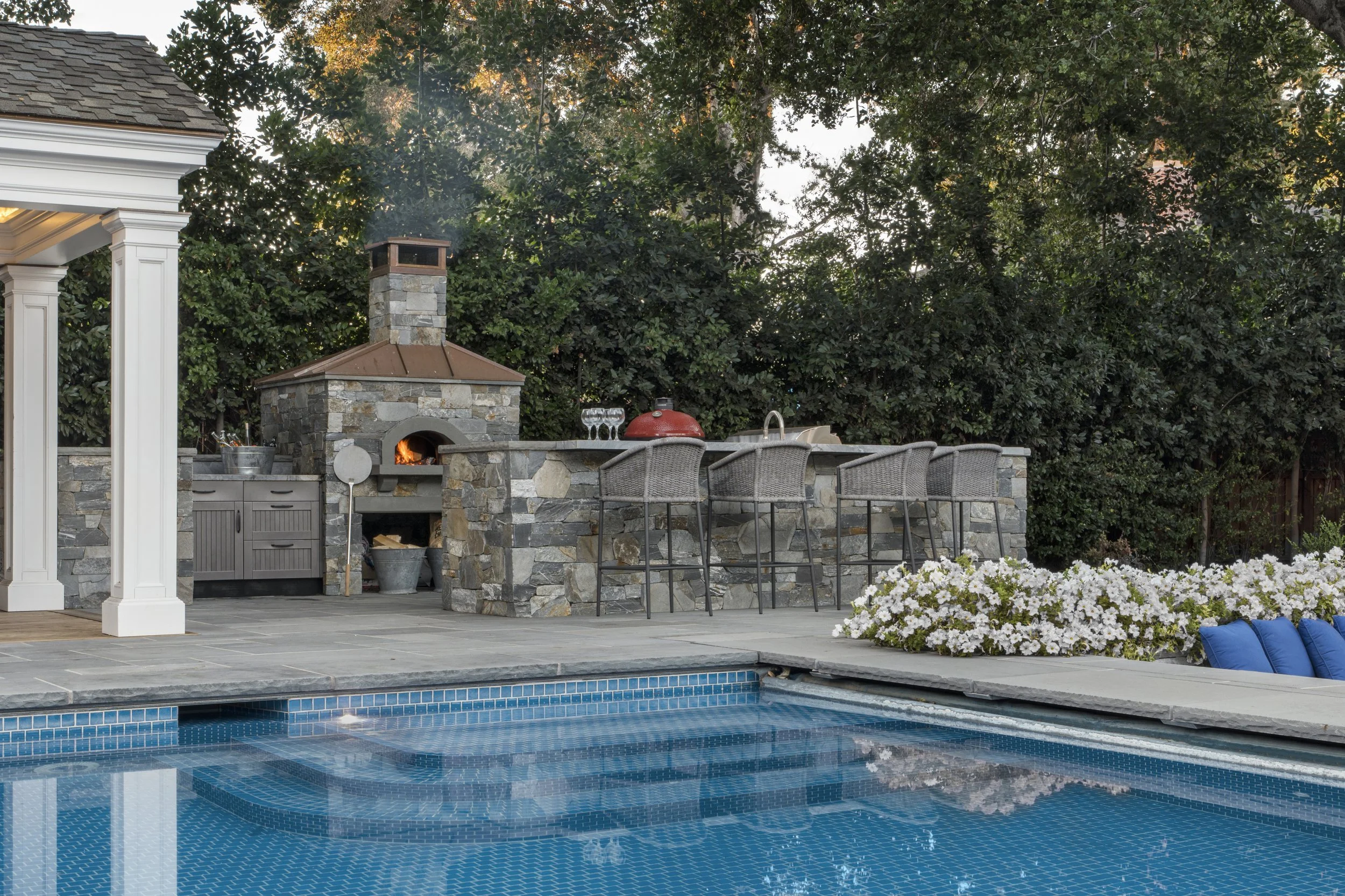 Backyard with a swimming pool, outdoor kitchen with stone fireplace, bar stools, and flowering bushes.