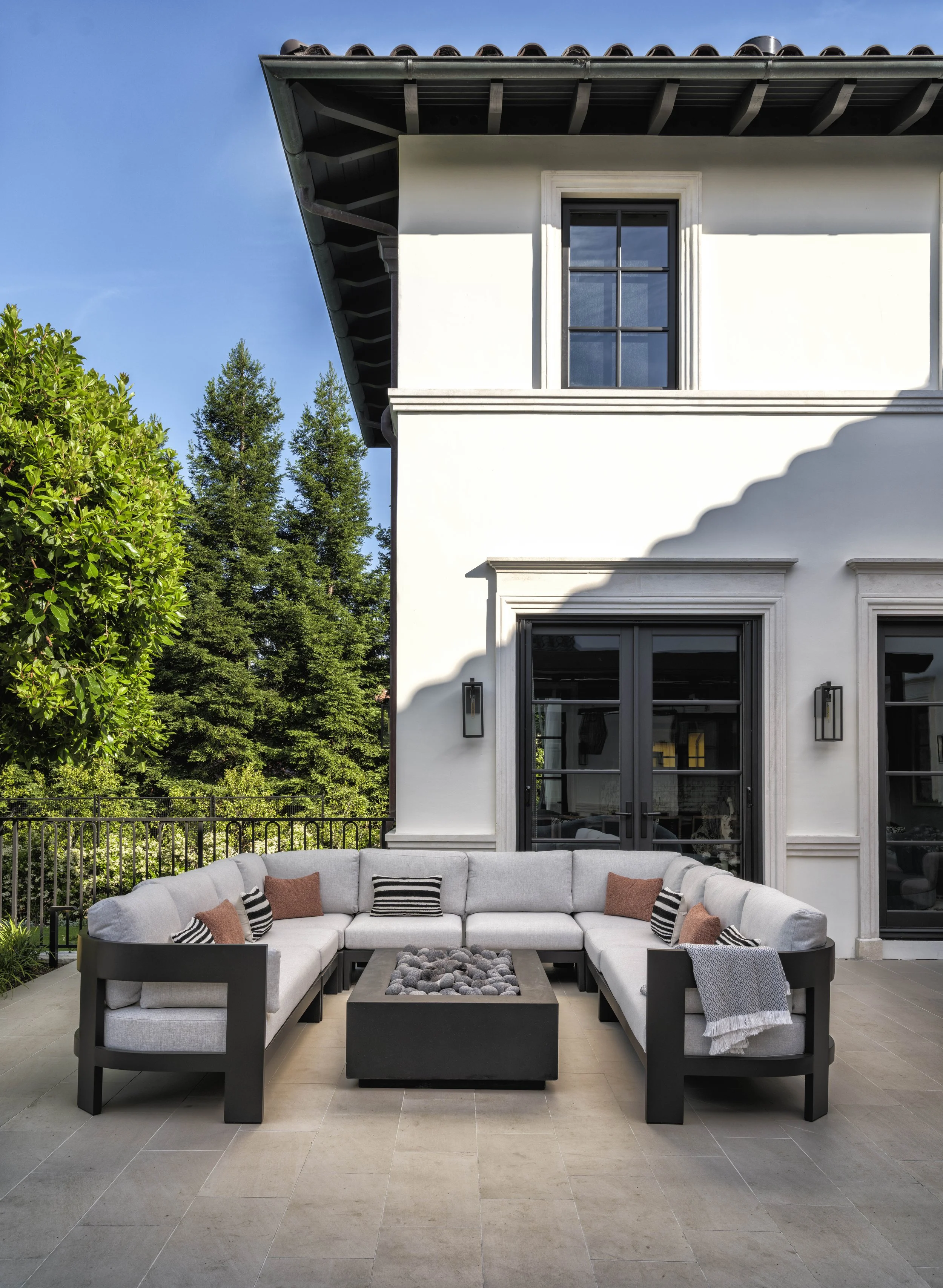 Modern outdoor patio with a large L-shaped gray sofa, decorative pillows, and a fire pit in the center. The patio is attached to a white house with black-framed glass doors and windows, surrounded by green trees under a blue sky.