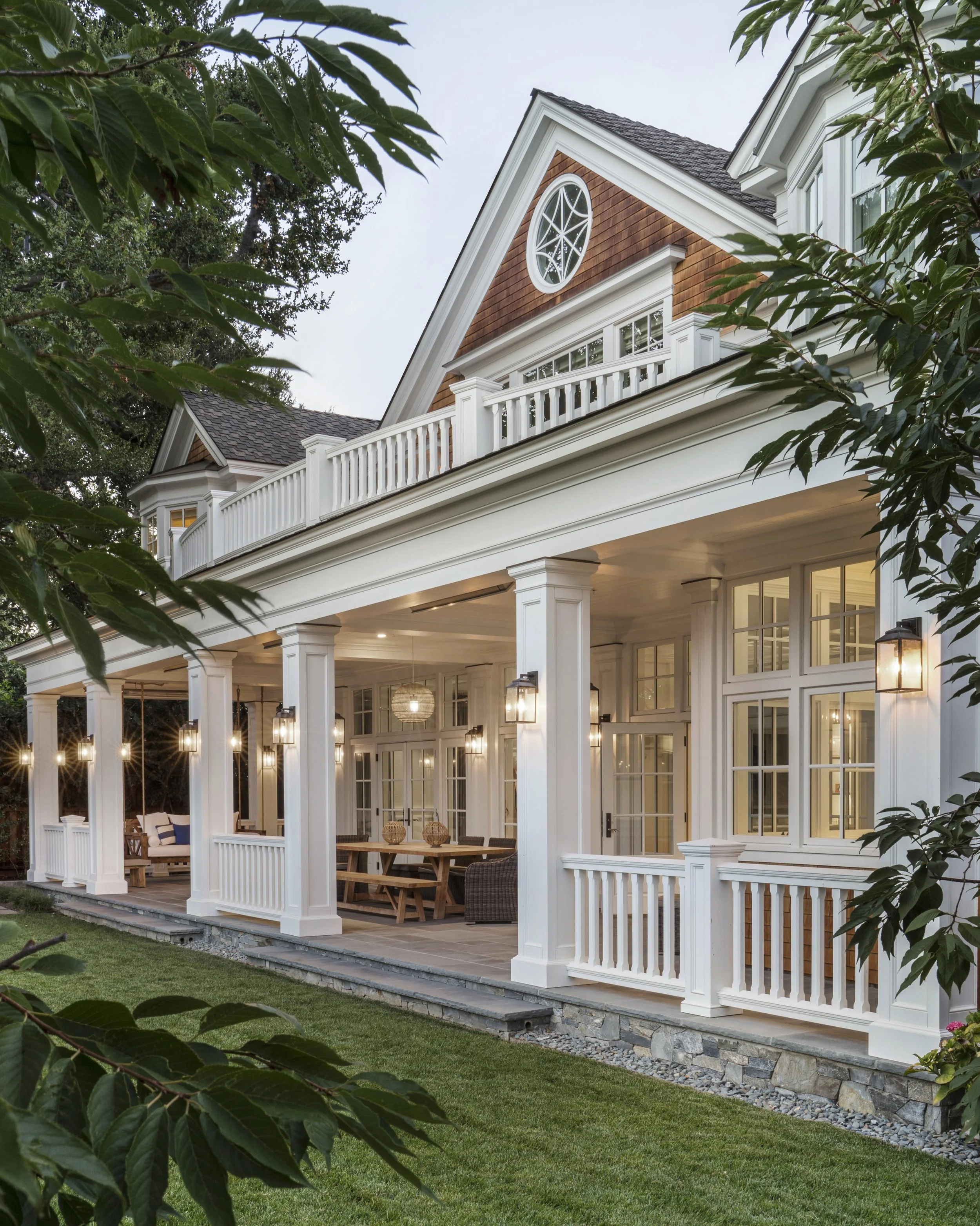 A large, elegant two-story house with white siding, multiple windows, and a covered patio with outdoor furniture and hanging pendant lights.