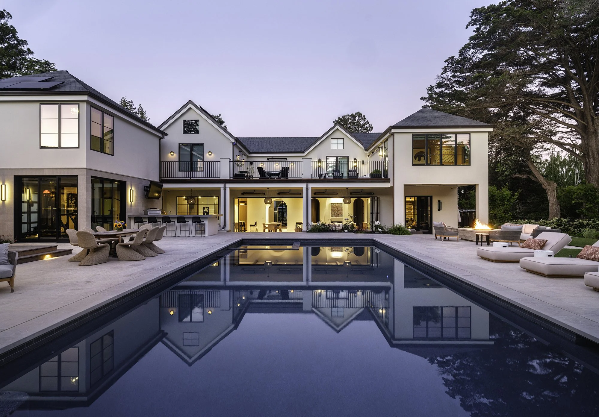 Luxury modern house with a large rectangular swimming pool in the backyard, outdoor seating, fire pit, and two floors with patio and balcony, surrounded by trees at dusk.