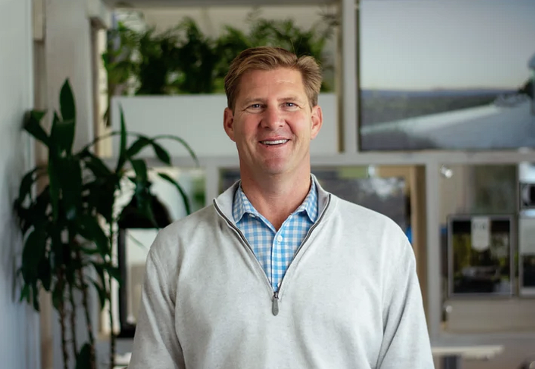 A man smiling in an indoor office setting with plants and monitors in the background.