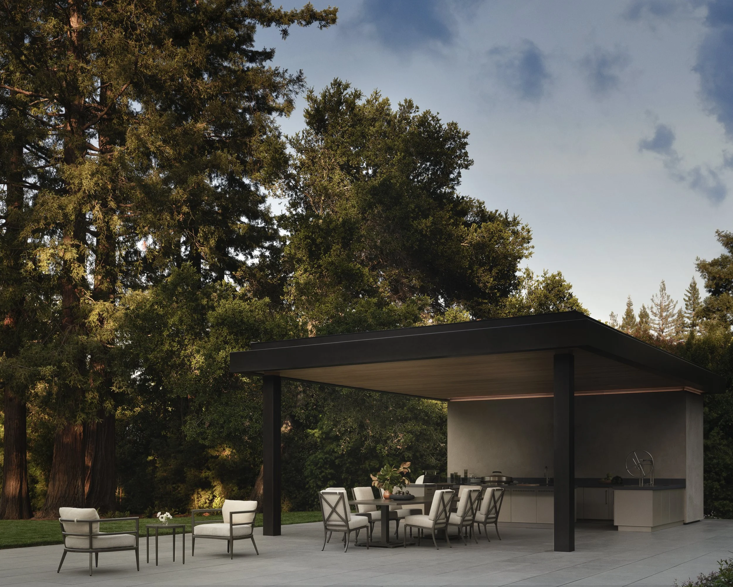 An outdoor patio with a modern open kitchen and dining area, surrounded by large trees under a partly cloudy sky, during sunset.