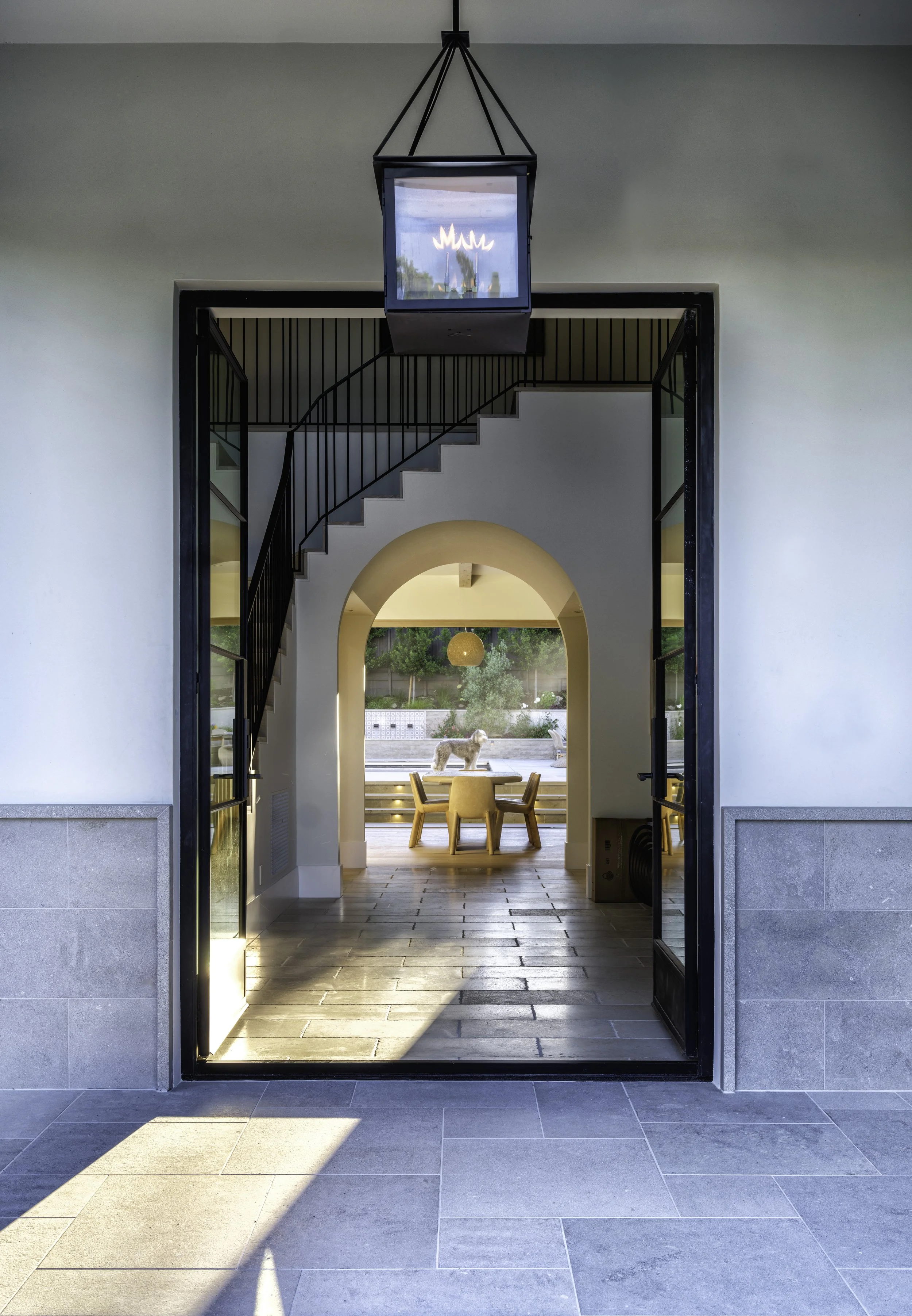 View through a black metal doorway into a modern dining area with wooden table and chairs, a large circular pendant light, and a dog outside on a patio with greenery in the background.