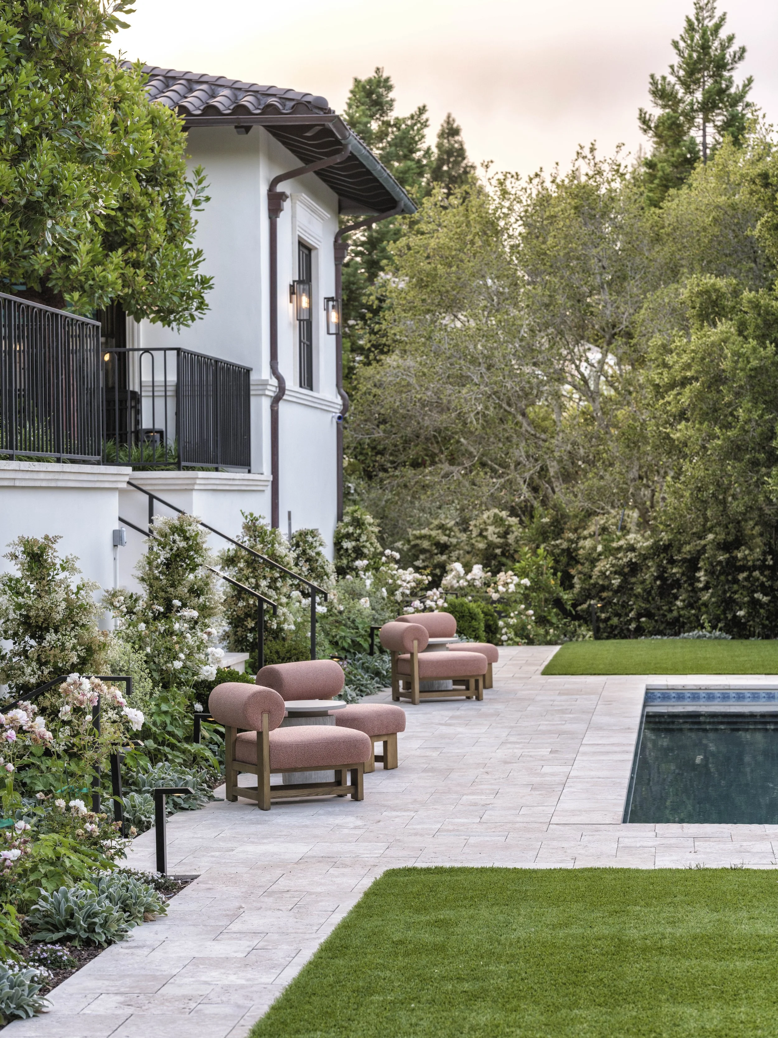 A backyard patio with a swimming pool, pink outdoor chairs, lush greenery, flowering bushes, and a white house with black railings.