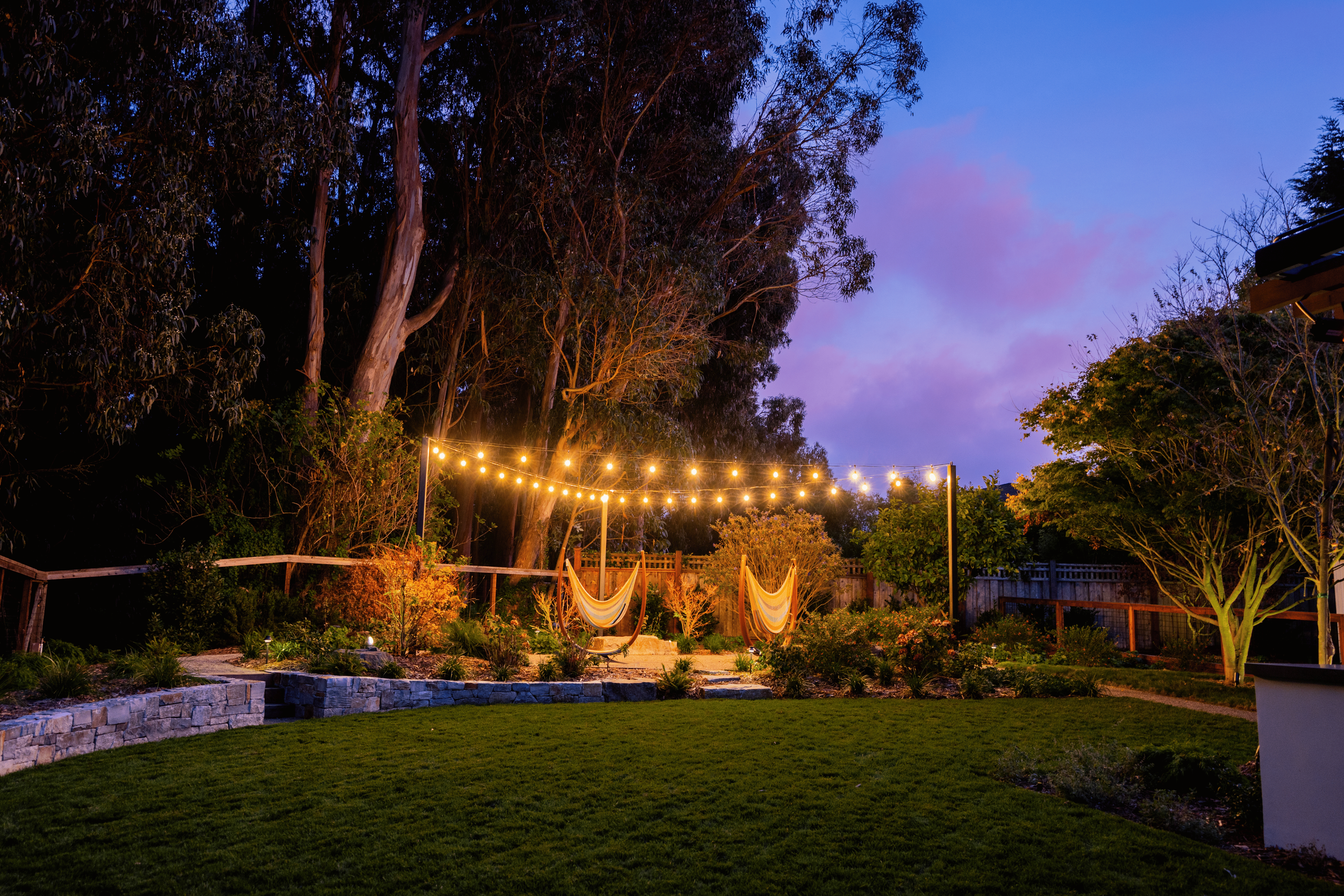 An outdoor backyard at dusk featuring a green lawn, stone-edged flower beds with small plants, and trees. String lights are hung overhead, casting a warm glow. Two hammocks are suspended in the background, surrounded by bushes and illuminated trees, 