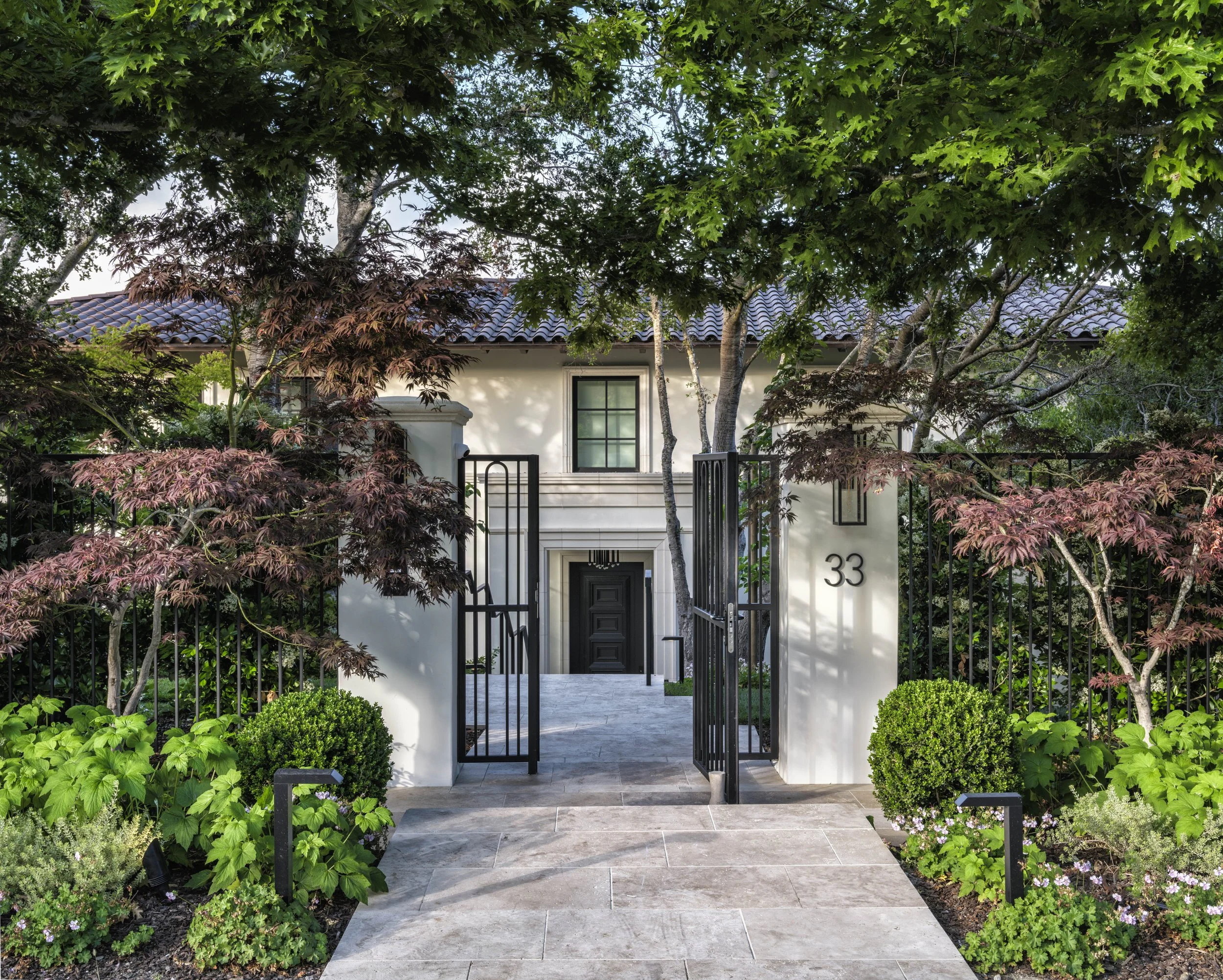Front view of a modern house with white walls, black door, and black gate, surrounded by lush greenery and garden plants with the house number 33 displayed on a white pillar.