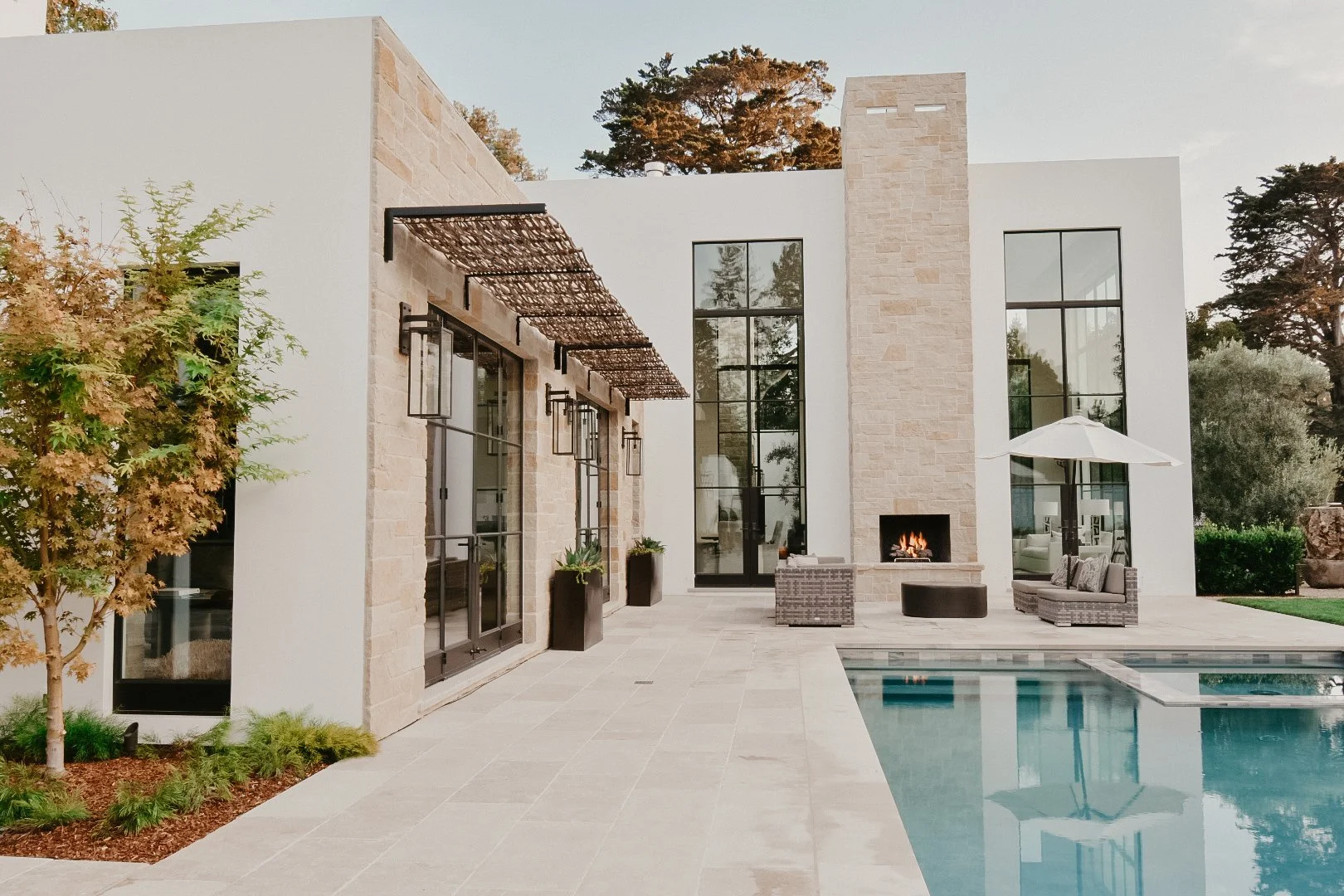 Modern backyard with a swimming pool, outdoor fireplace, patio seating, and large windows reflecting trees, with a tree and greenery on the left and trees in the background.