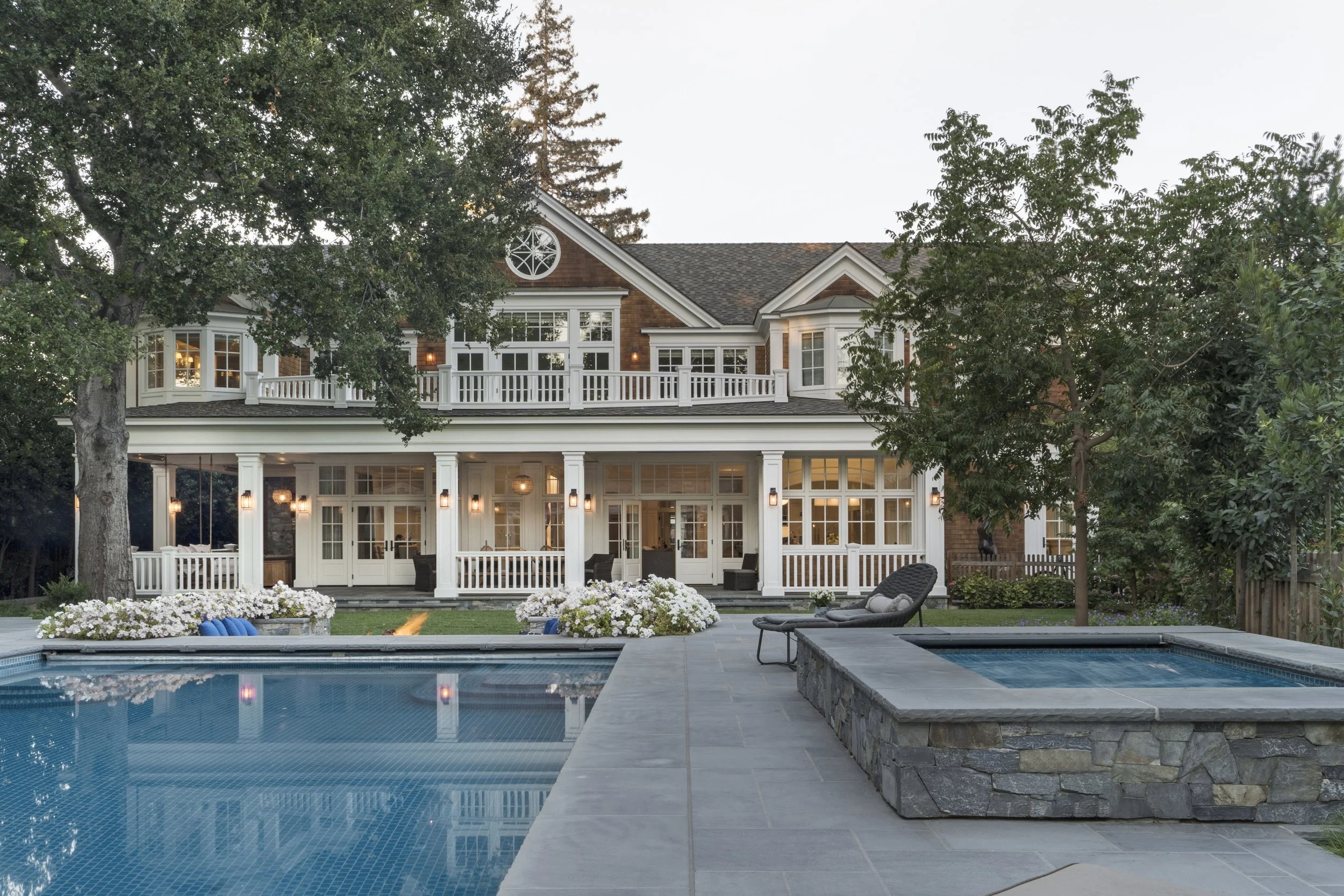 Large house with a backyard pool and hot tub, surrounded by trees and outdoor furniture, during dusk with interior lights on.