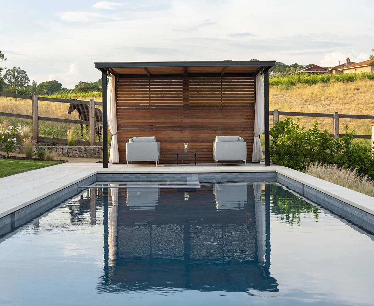 An outdoor backyard swimming pool with a wooden cabana that has two light gray armchairs and a small table with a drink. The pool reflects the cabana and the sky.