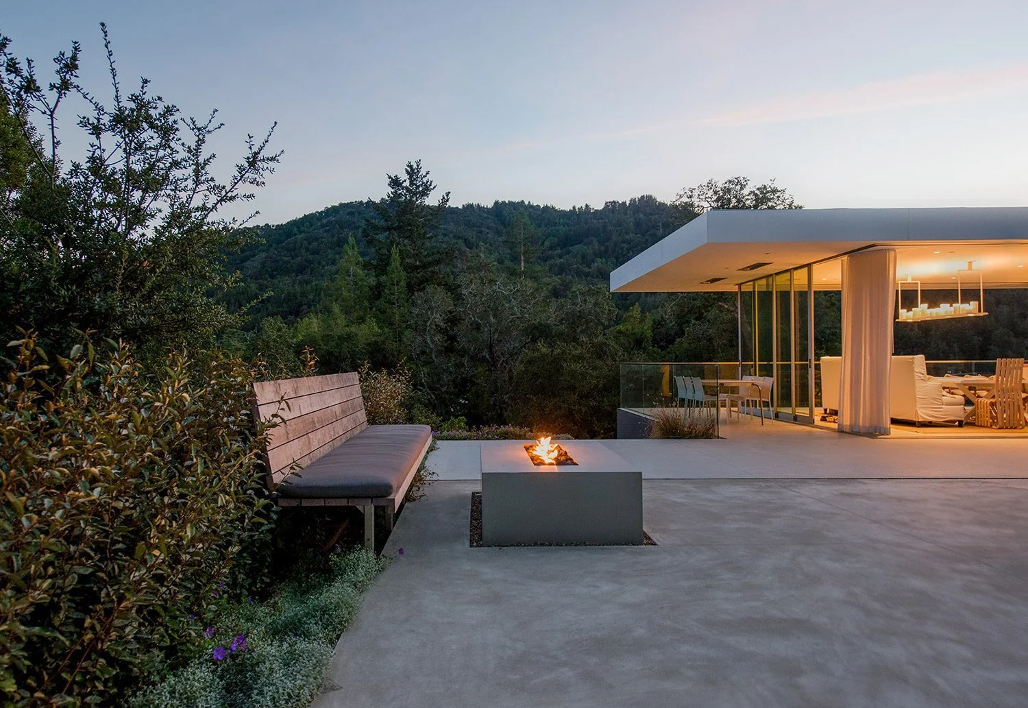 Modern house with an outdoor patio featuring a fire pit, a wooden bench, green bushes with flowers, surrounded by forested hills at dusk.