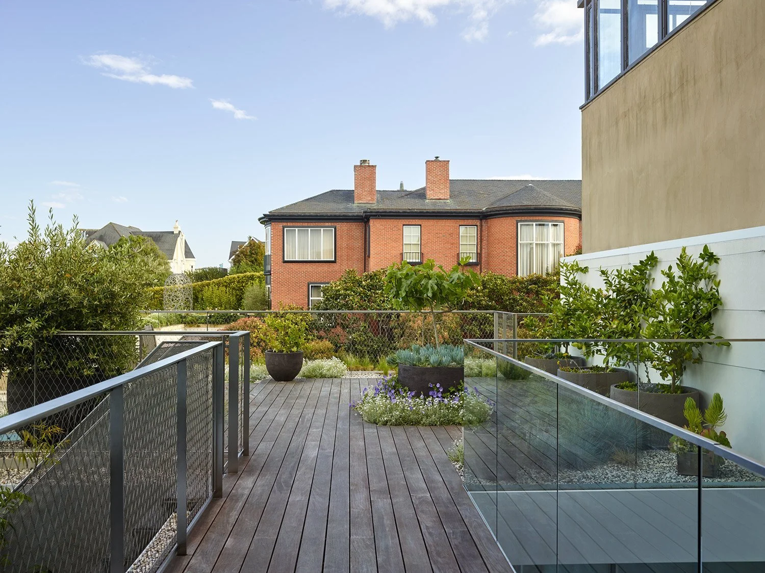 View of a modern balcony garden with wooden flooring, potted plants, shrubs, and trees, overlooking a brick residential building under a partly cloudy sky.
