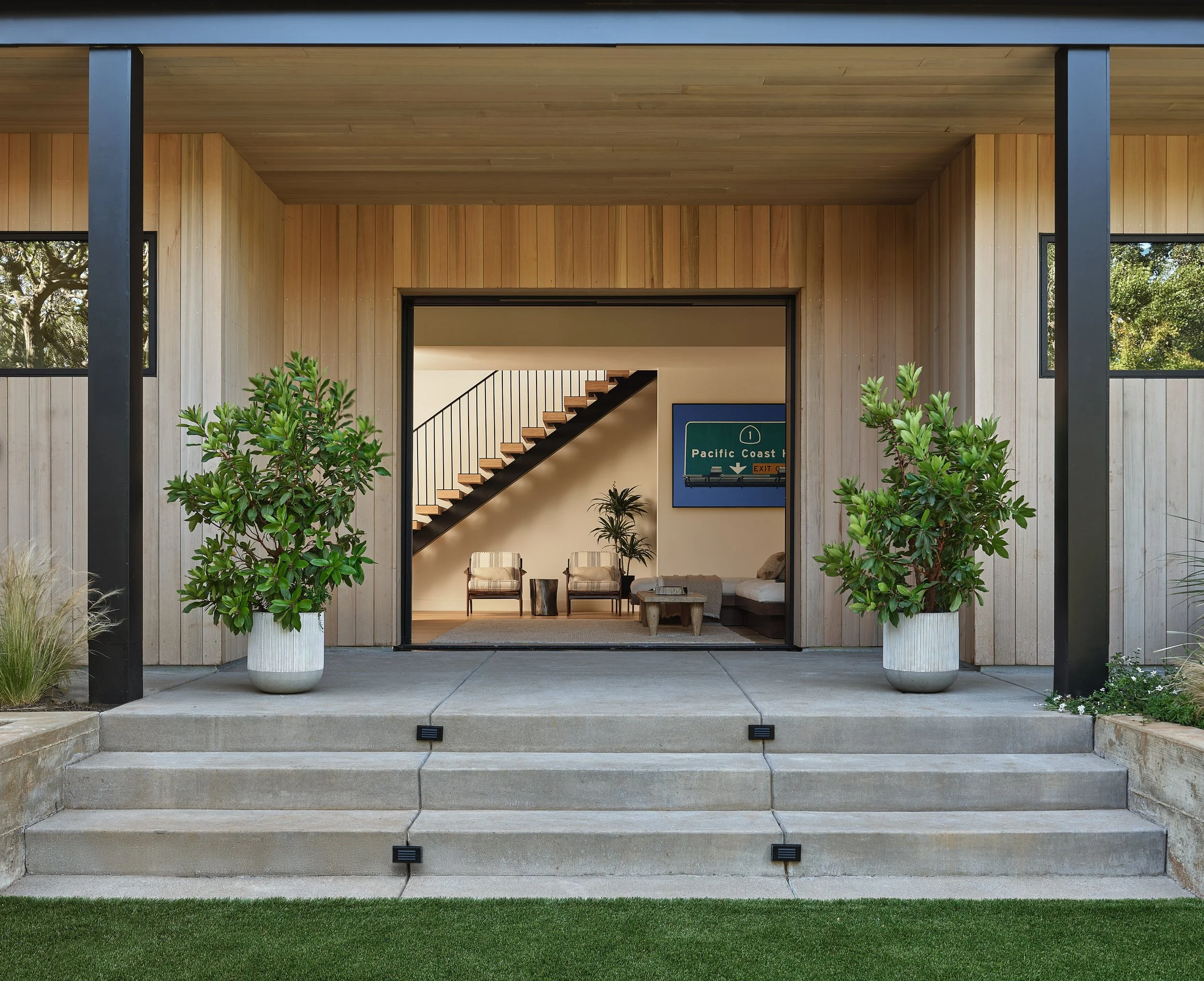 Modern house entrance with beige wooden siding, large potted green bushes on concrete steps, and interior living space visible through open sliding doors, featuring a staircase, seating area, and a framed sign on the wall.