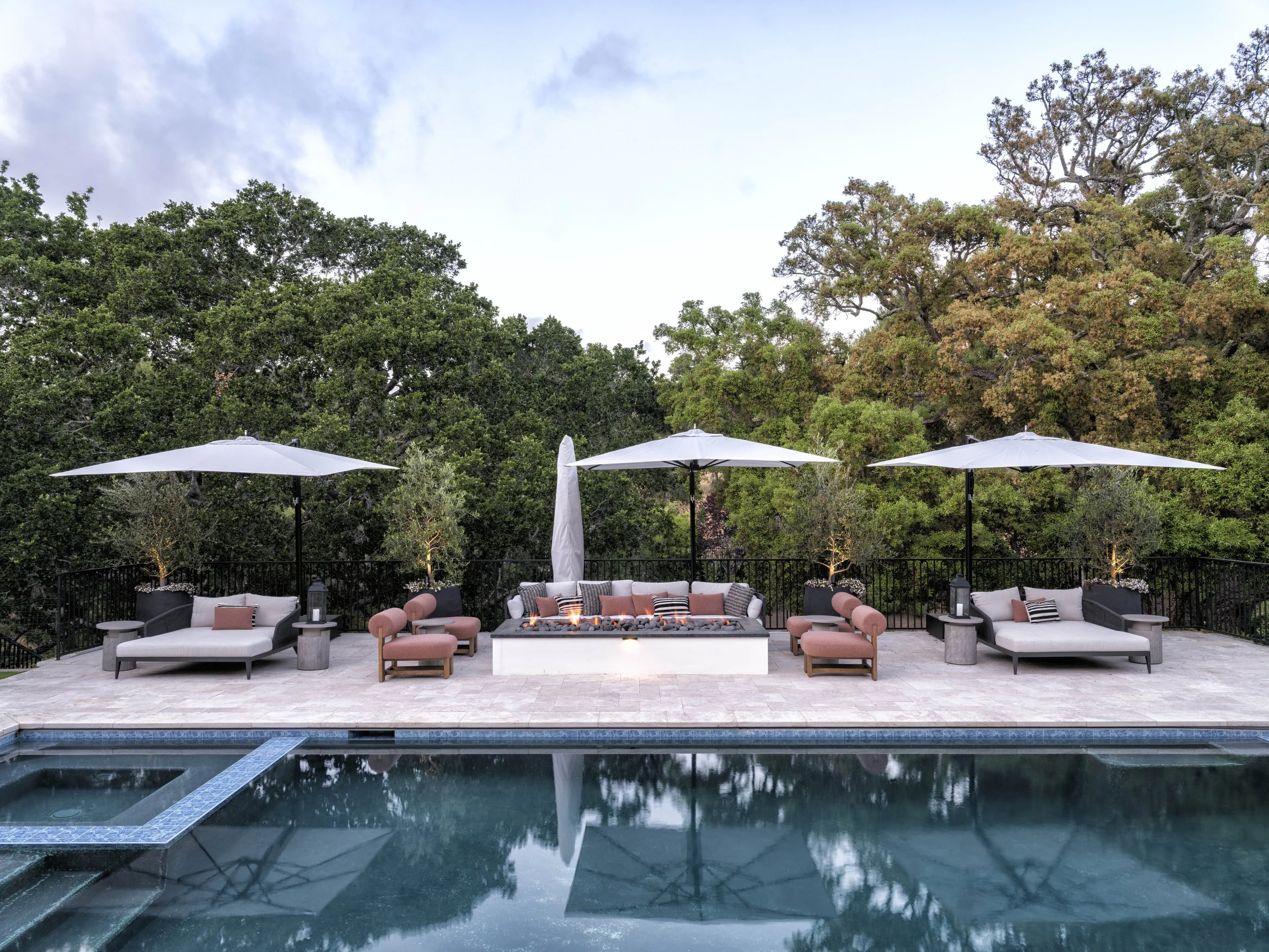 Poolside outdoor seating area with three large white umbrellas, lounge chairs, and a fire pit with surrounding chairs, set against lush green trees