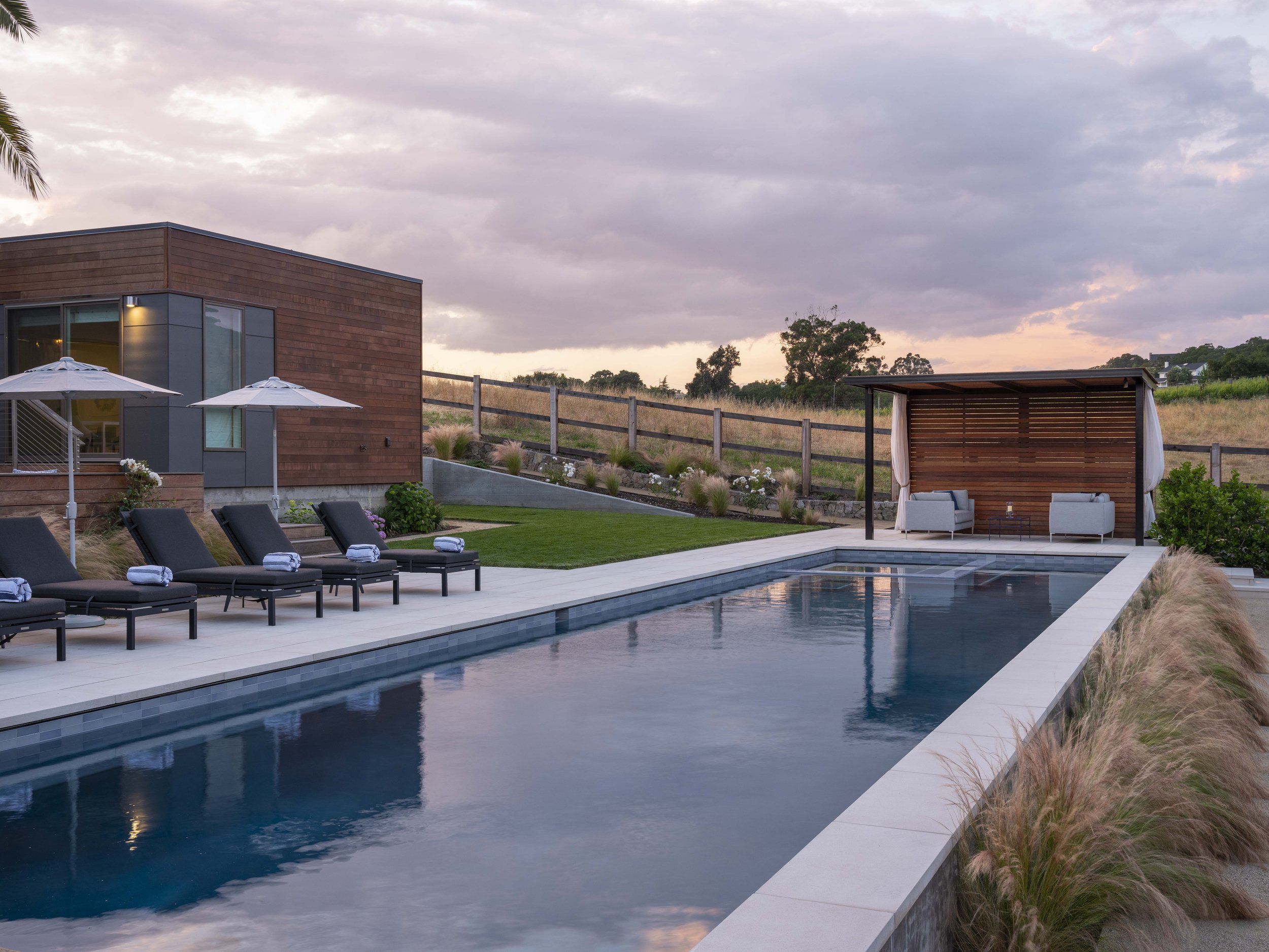Luxury backyard with rectangular swimming pool, lounge chairs with rolled towels, umbrellas, a modern house, and a shaded seating area with two couches, set against a sunset sky and grassy hillside.
