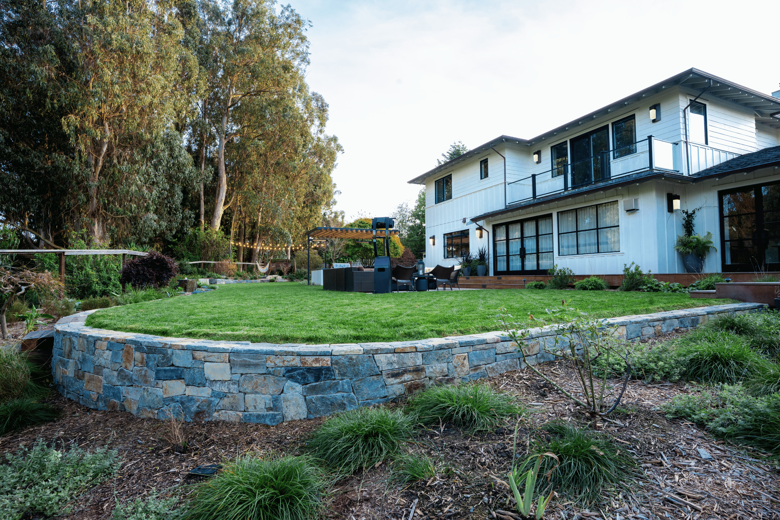 A backyard with a lush green lawn, surrounded by plants and trees. There is a stone retaining wall in the foreground and a white two-story house with large windows and a balcony in the background. Outdoor seating and string lights are visible with a 