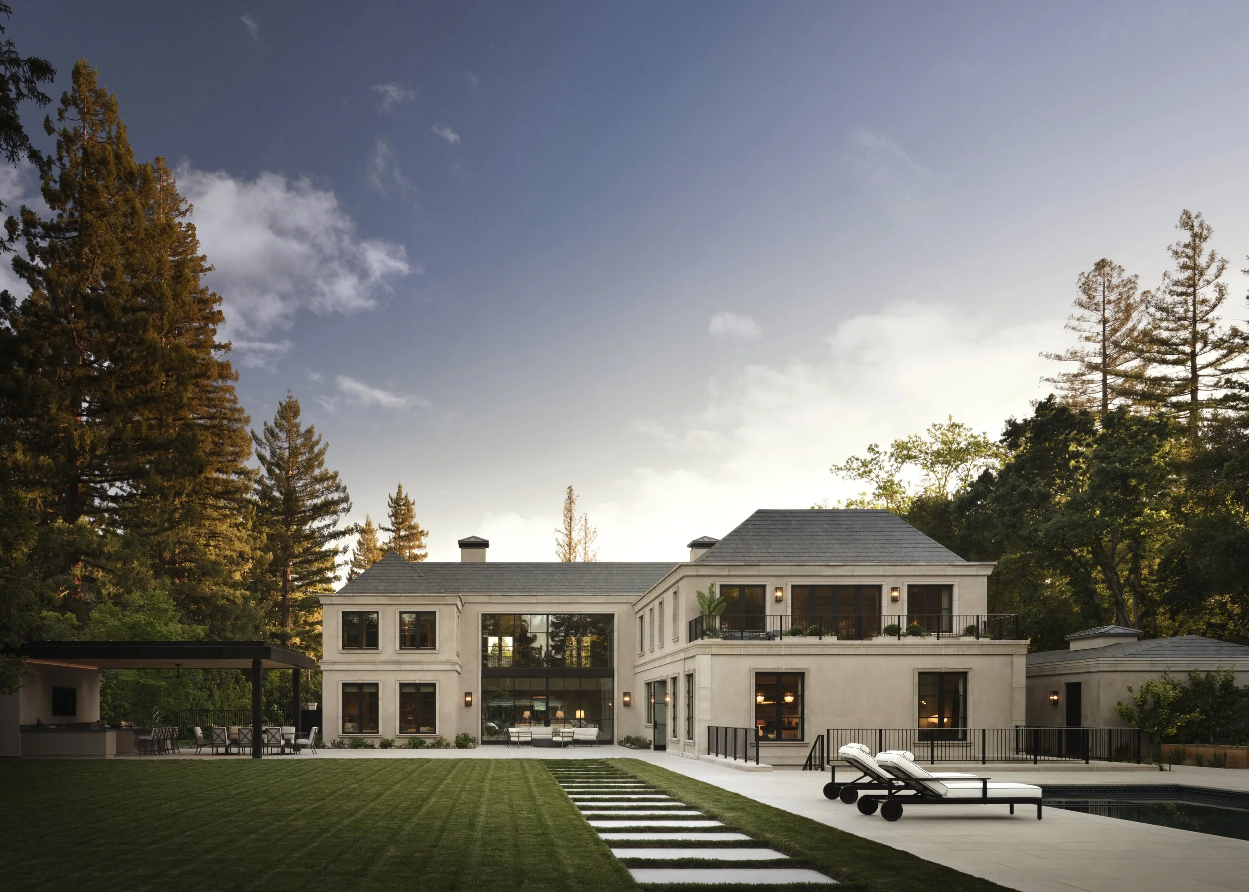 Modern two-story house with large windows, balcony, outdoor patio, and pool area, surrounded by tall trees, during sunset.