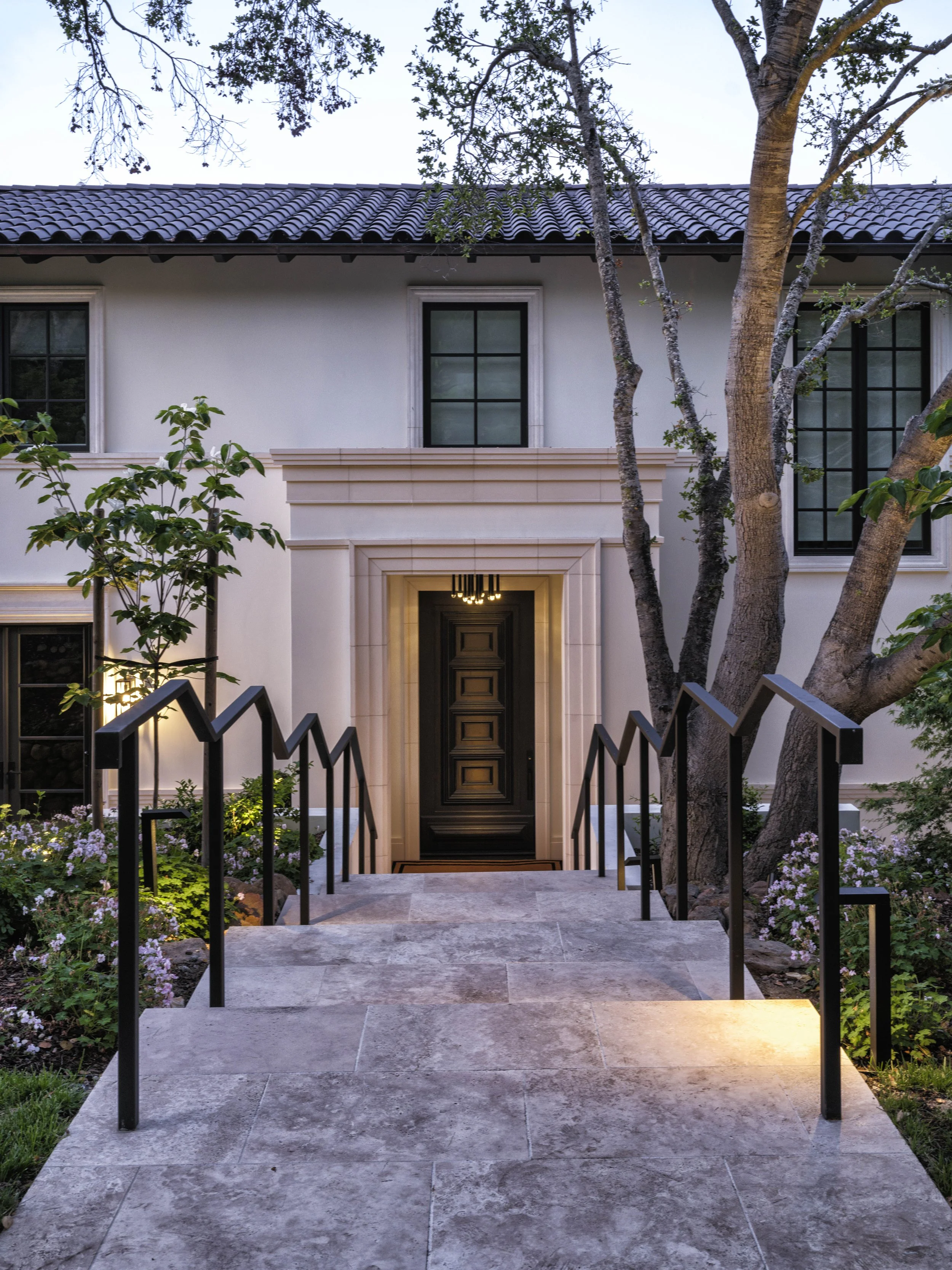 Front entrance of a modern house with a staircase leading to the door, surrounded by trees and plants, illuminated at dusk.