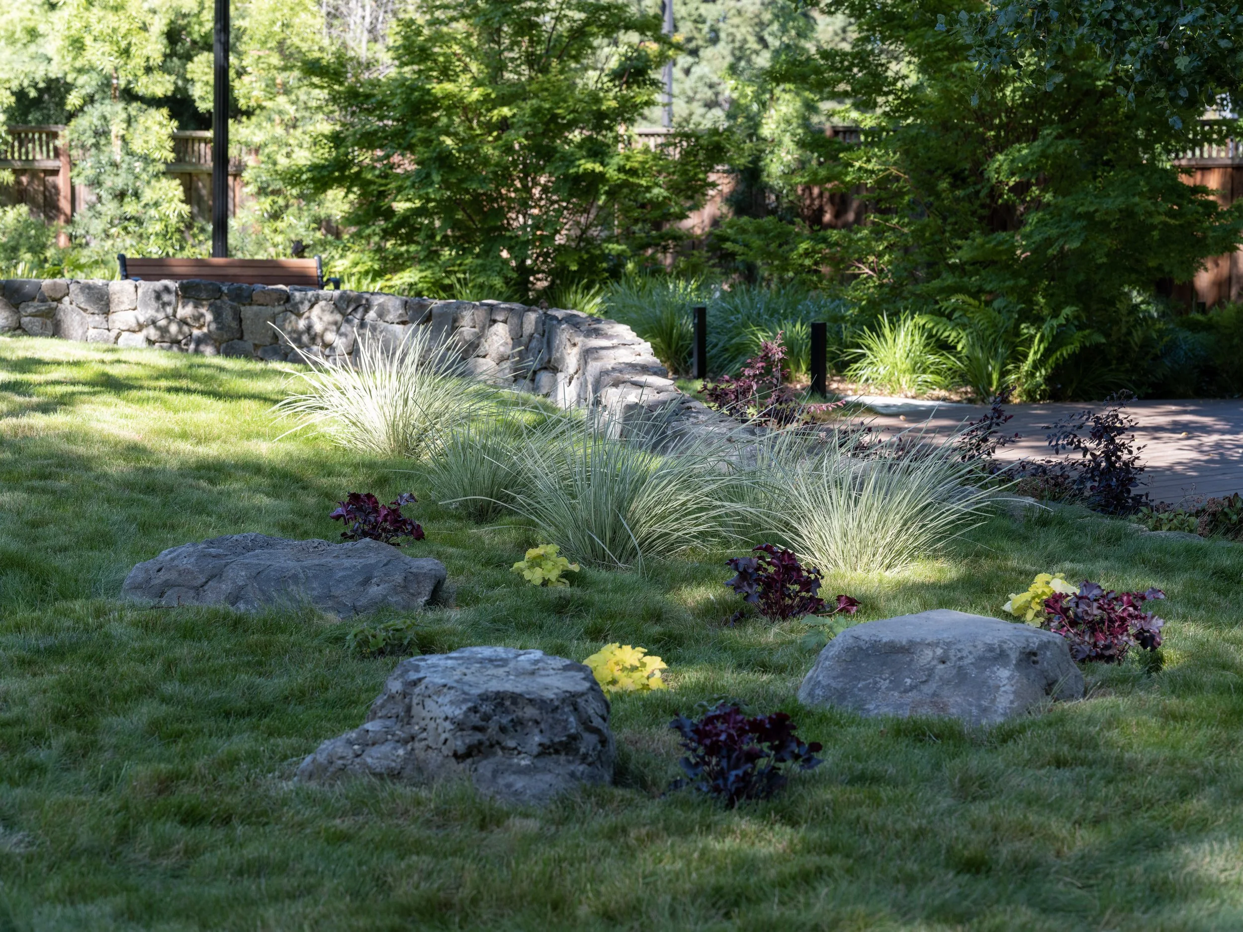 A landscaped garden with grass, rocks, ornamental plants, and a stone wall, shaded by trees and illuminated by sunlight.