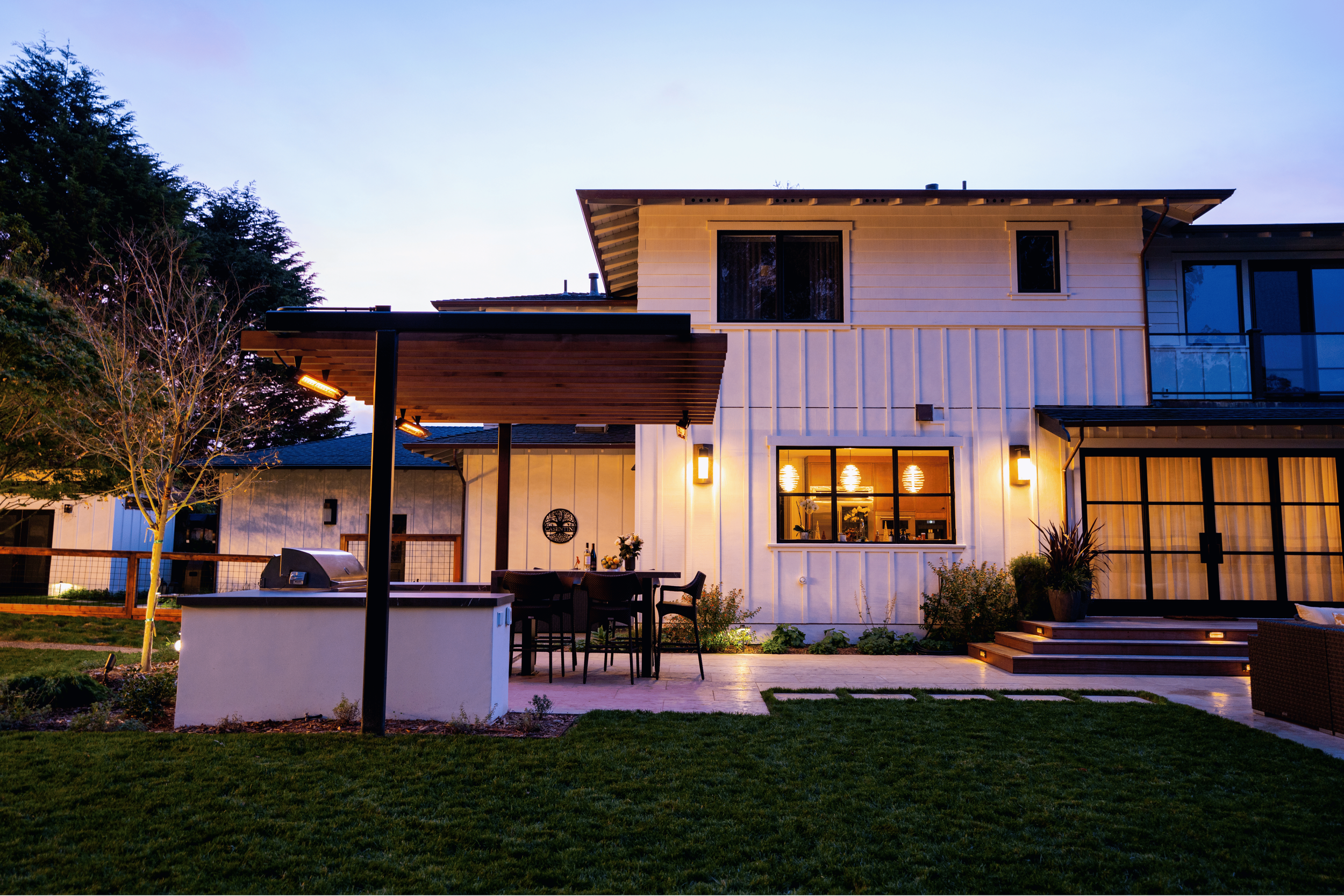 Modern two-story house with outdoor patio and lawn, illuminated by exterior lights during dusk.