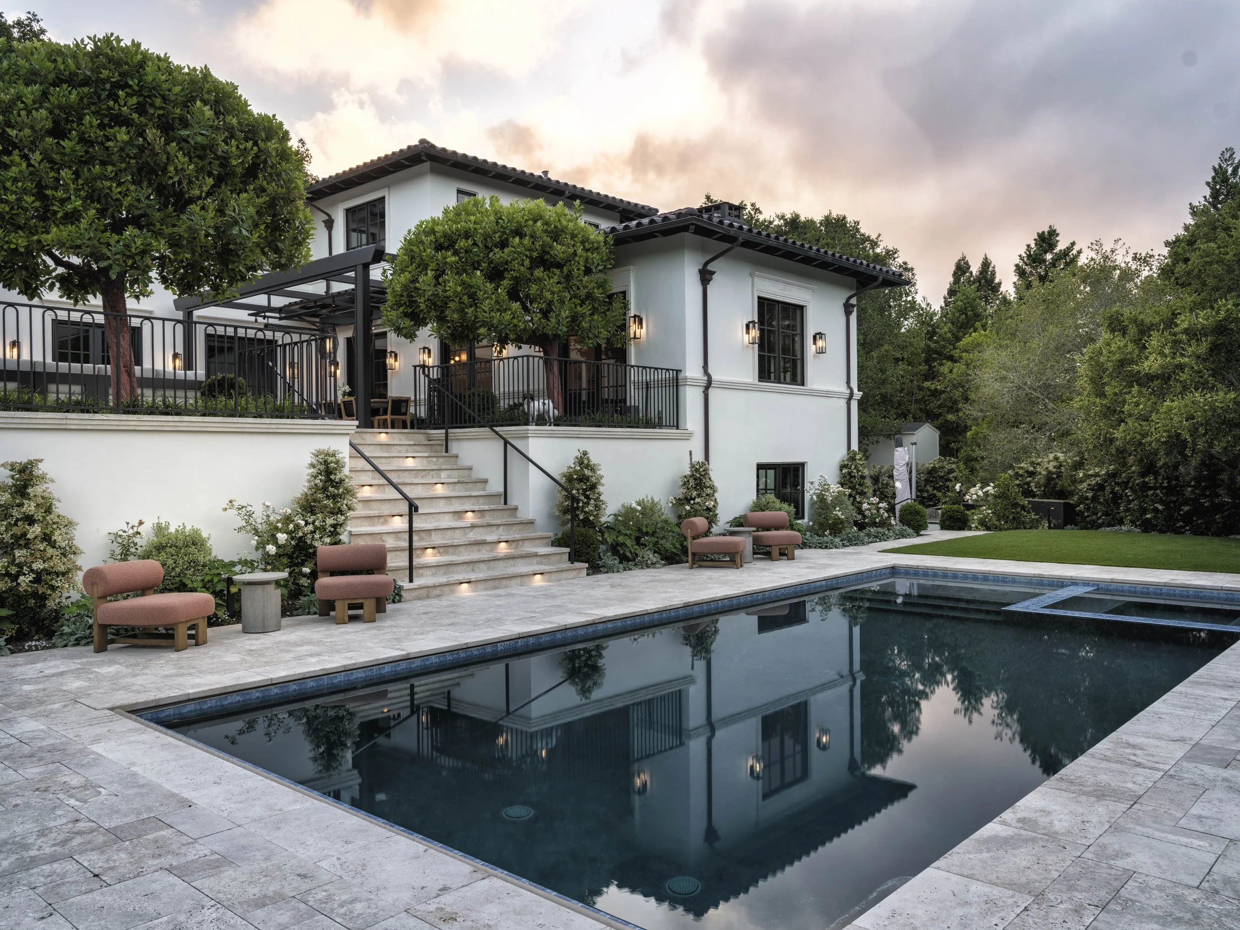 Luxury house with a swimming pool, outdoor lounge area, stairs, manicured garden, and trees at sunset.