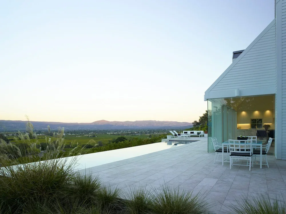Modern house with outdoor patio, dining set, and pool overlooking a valley and distant mountains during sunset.