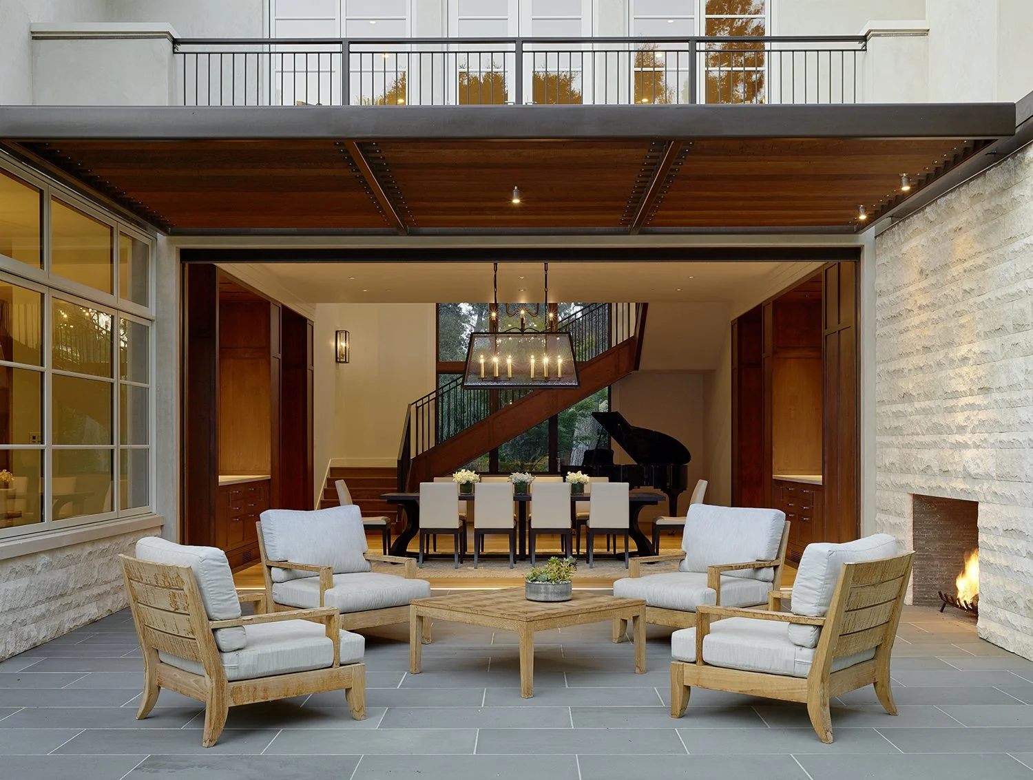 Outdoor patio with four wooden armchairs with cushions arranged around a low wooden table, a fireplace on the right, and an open indoor area featuring a dining table with chairs, a black grand piano, and a staircase in the background.