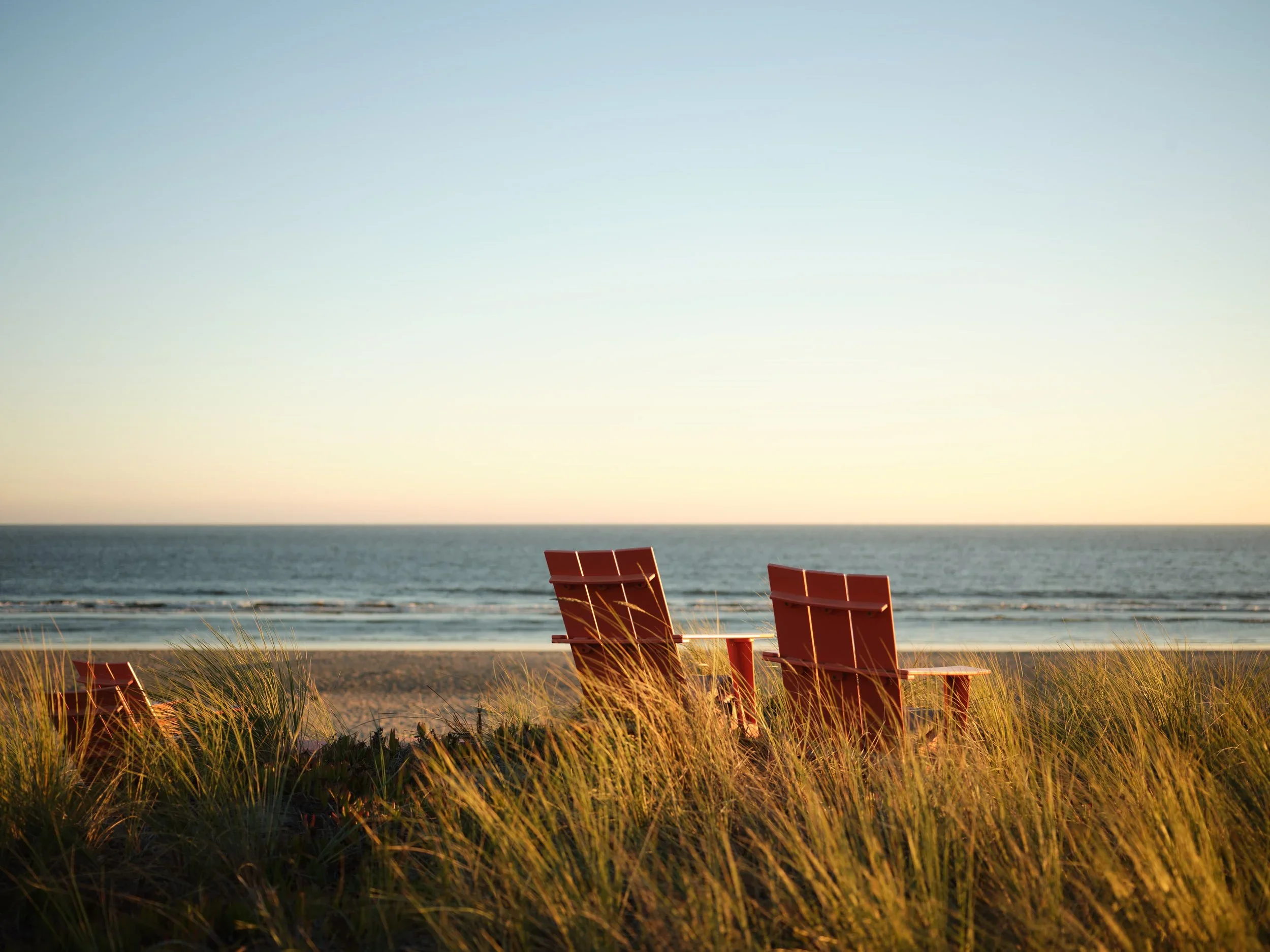 Three red beach chairs on the sand among tall beach grass facing the ocean at sunset.
