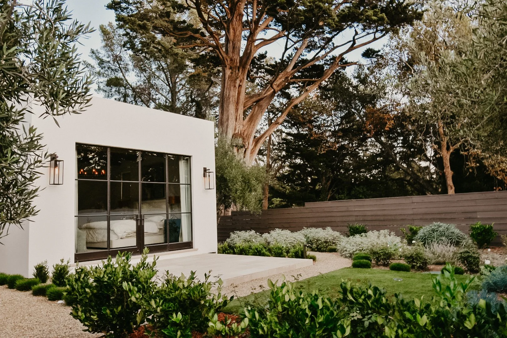A modern white house with large glass doors, outdoor lanterns, and a landscaped yard with various green shrubs and trees.