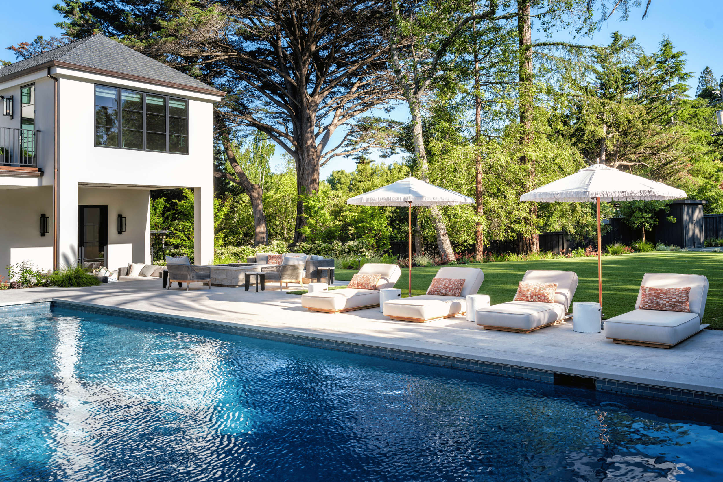 Luxury backyard with a swimming pool, lounge chairs with umbrellas, and a modern two-story house surrounded by trees and greenery.