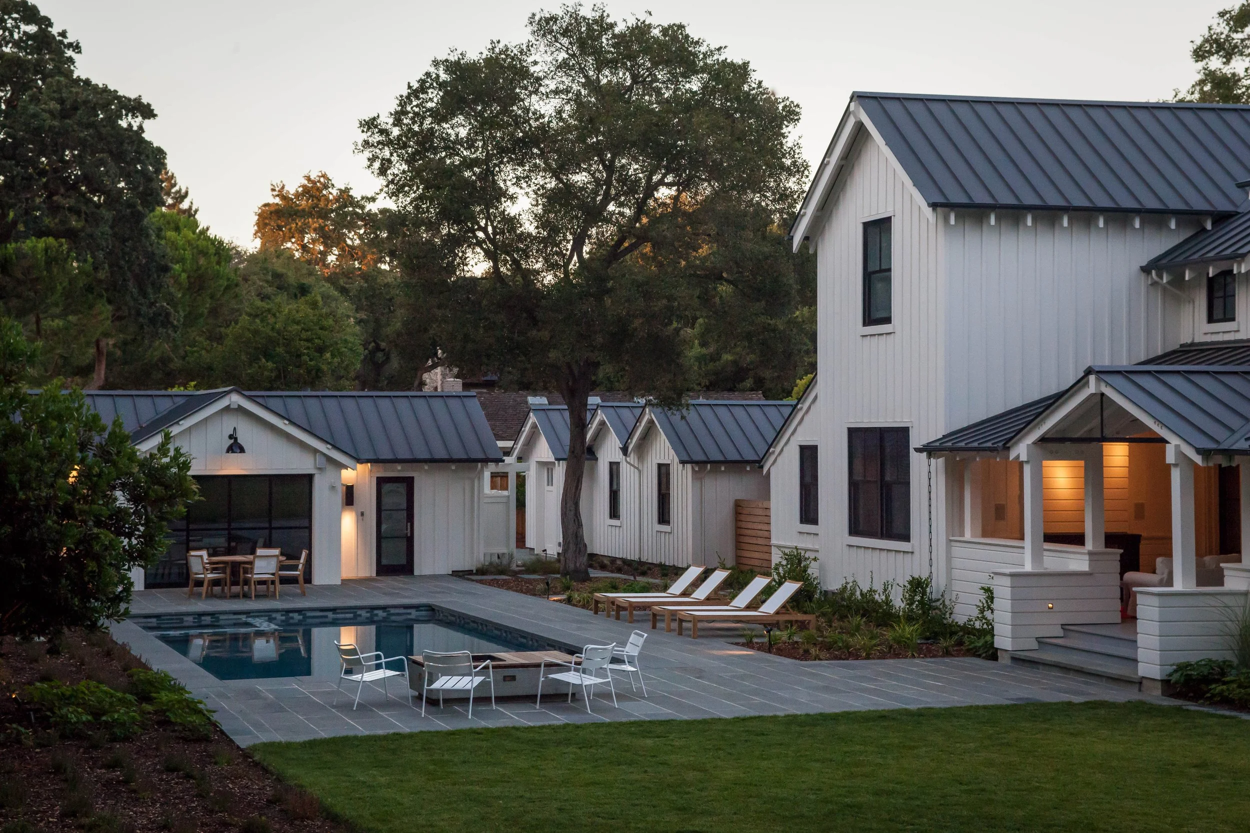 A backyard view of modern white houses with black roofs, a swimming pool with lounge chairs, outdoor dining area, and trees during sunset.