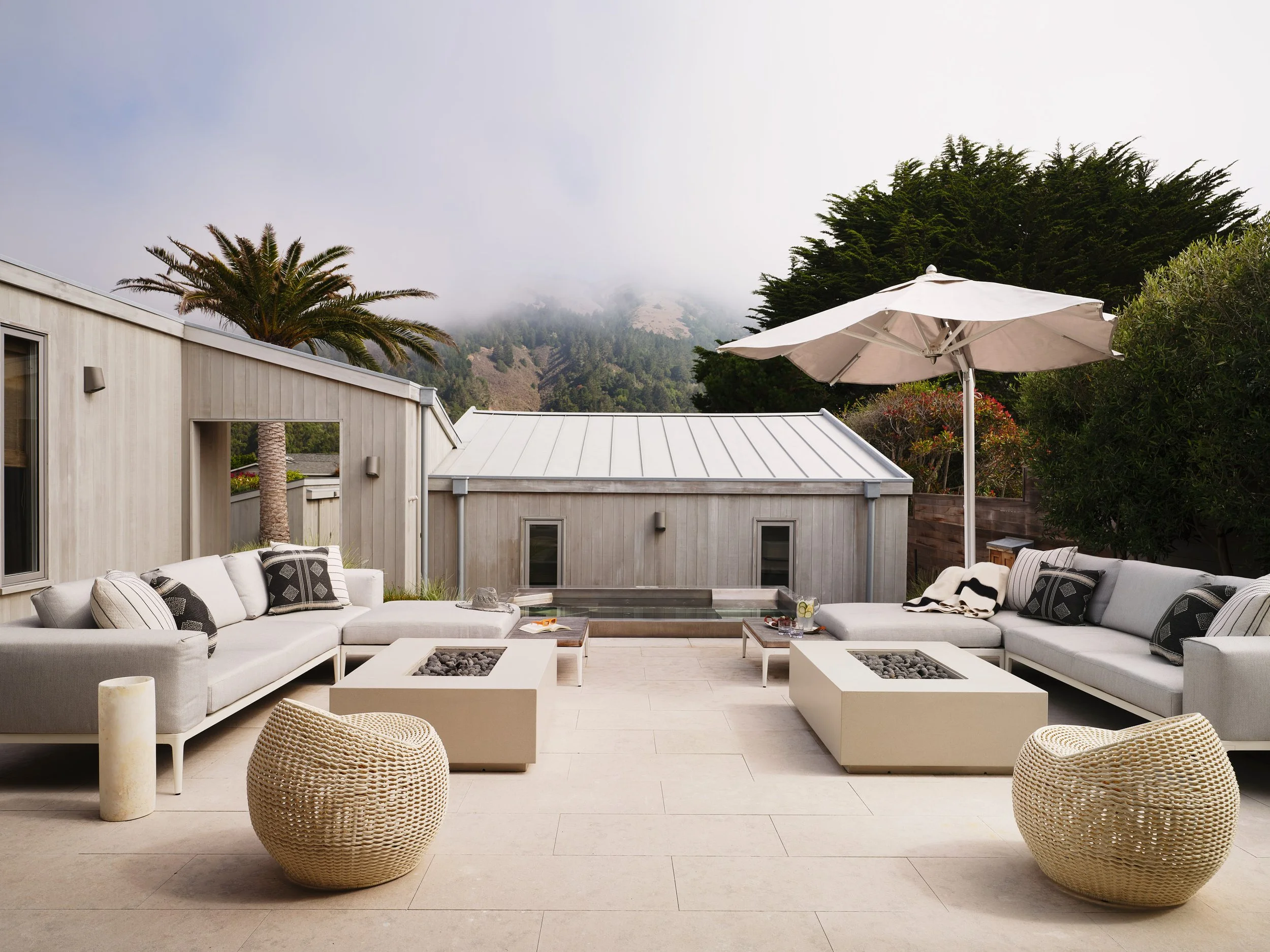An outdoor patio with modern sofas, a large umbrella, fire pits, and mountainous scenery in the background.