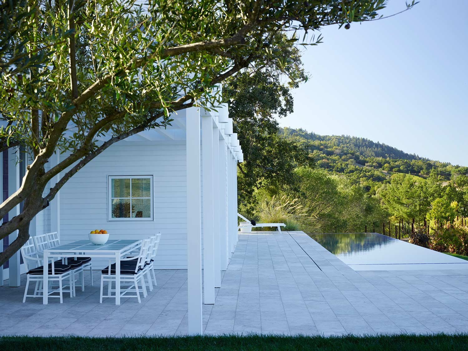White outdoor patio with dining table and chairs, bowl of fruit, and a view of green rolling hills and trees in the background.