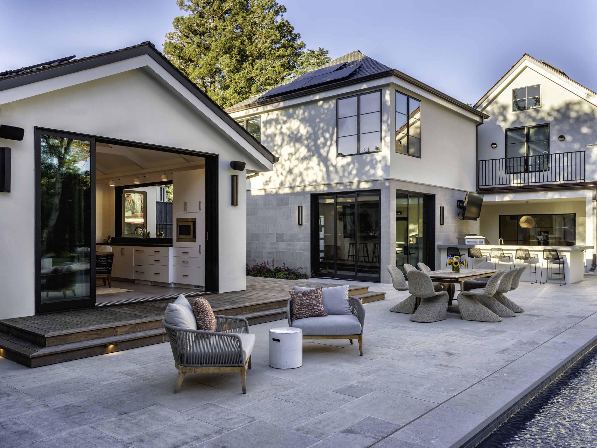 Modern backyard outdoor living space with white house, patio furniture, dining table with chairs, outdoor kitchen, and a swimming pool.