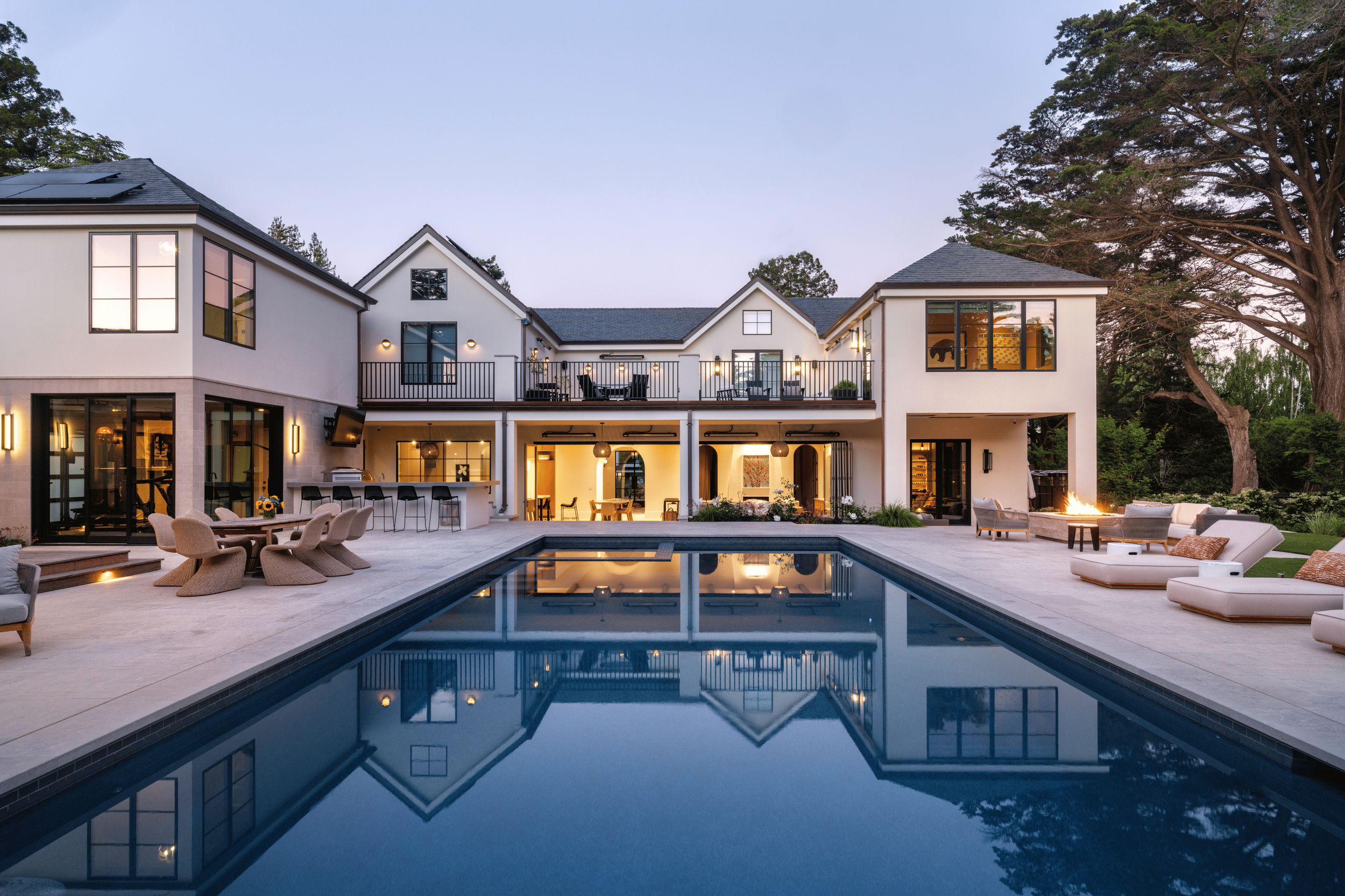 Modern two-story house with large windows, spacious balcony, and outdoor seating area around a swimming pool at dusk.
