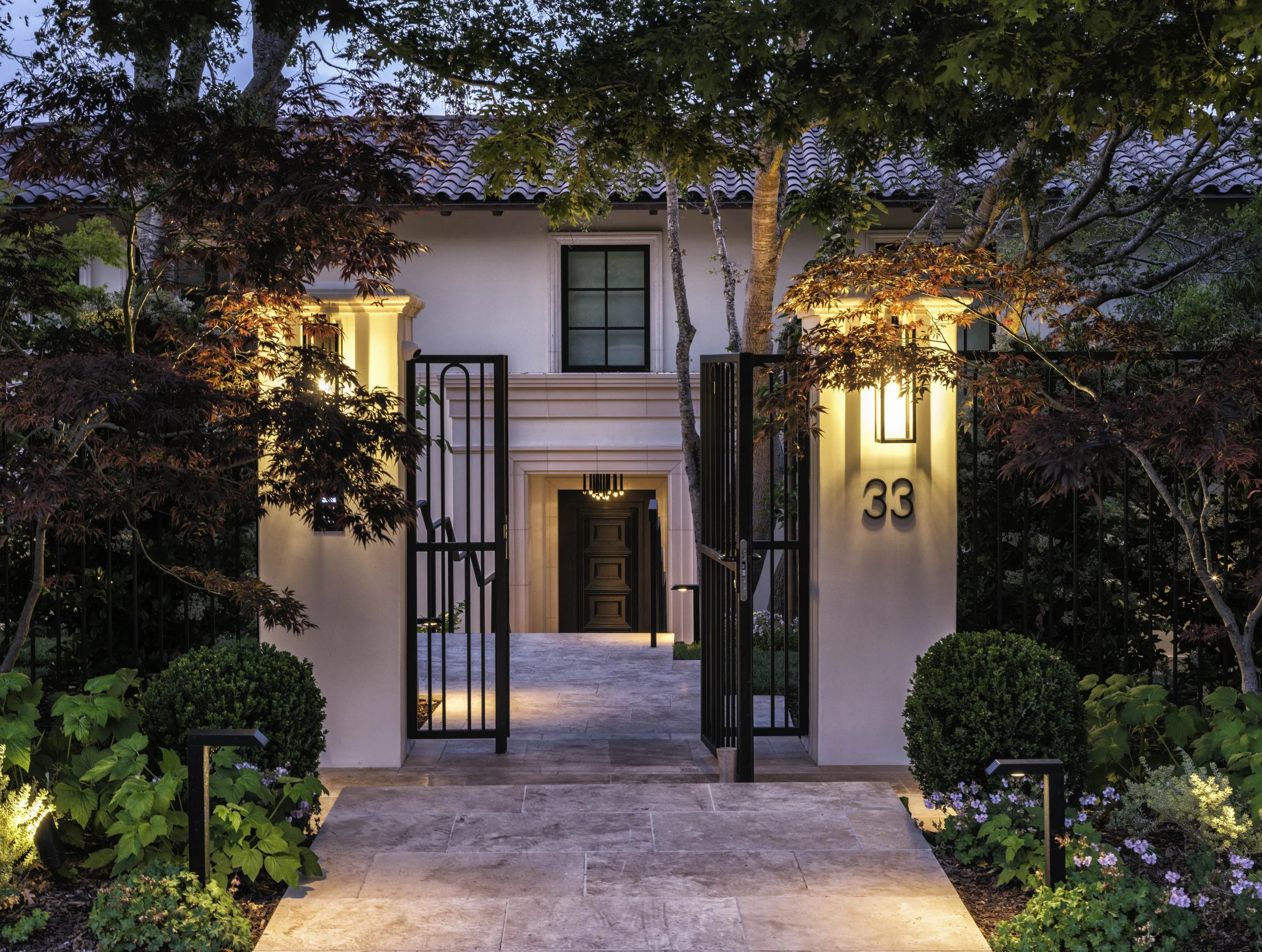Front view of a modern house with a gated entrance, illuminated exterior lights, surrounded by lush greenery and trees, with house number 33 displayed on the wall.