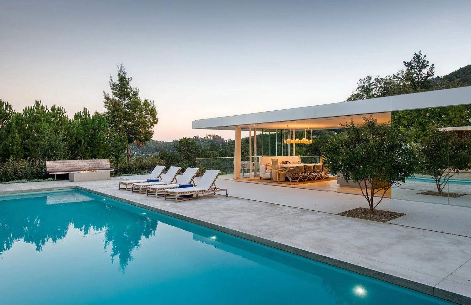 Modern outdoor swimming pool area with lounge chairs, a fire pit, and an open-concept patio with lighting, surrounded by trees and hills at dusk.