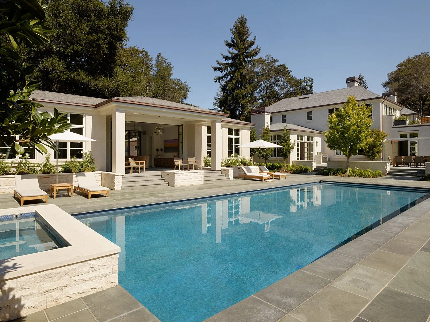 A luxurious backyard with a large swimming pool, poolside loungers with umbrellas, a modern house with large windows, trees, and a porch area in a sunny day.