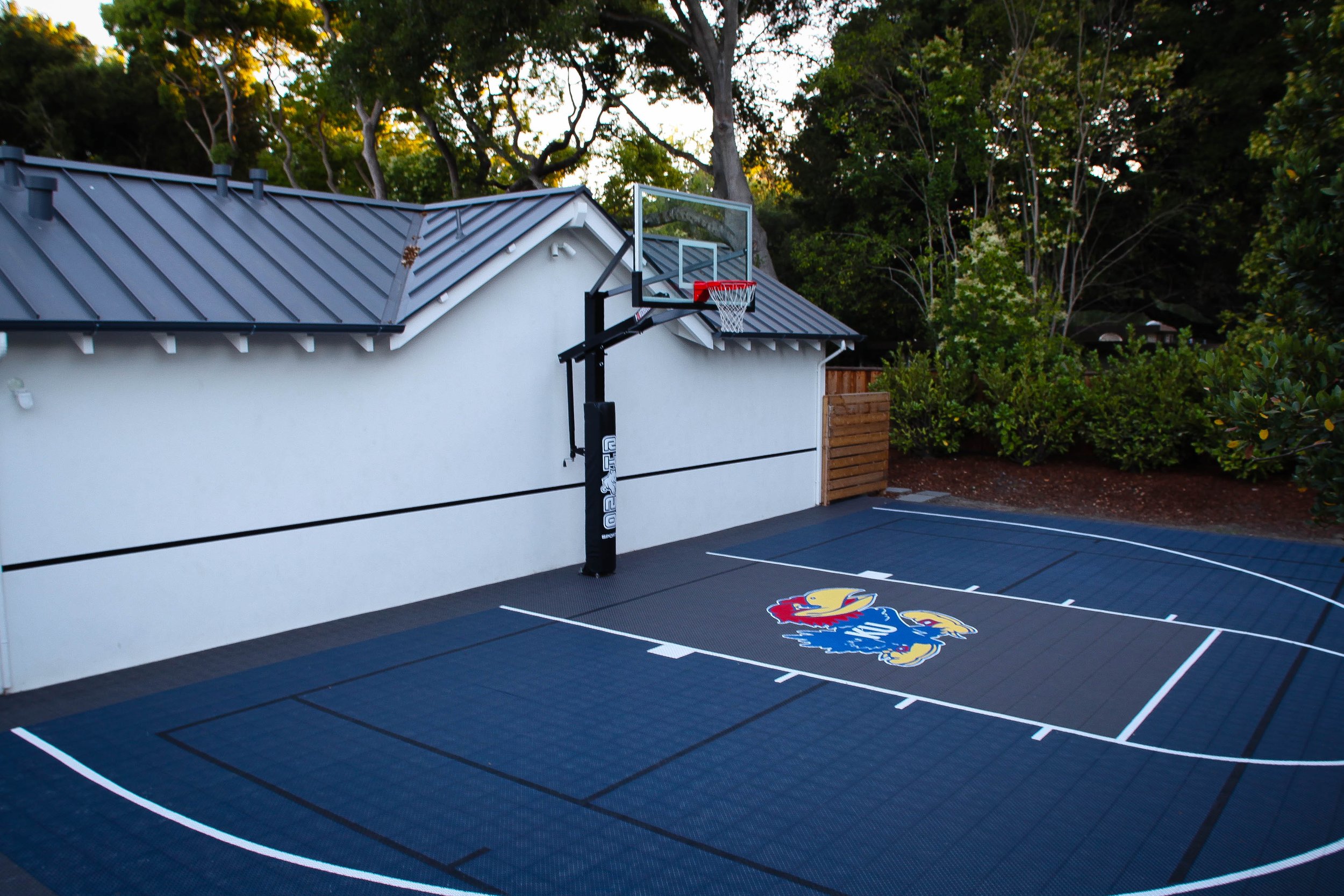 An outdoor basketball court with black surface, white lines, and a Kansas Jayhawks logo at the center. A basketball hoop with backboard is mounted on the wall beside a white building, with trees and bushes in the background.