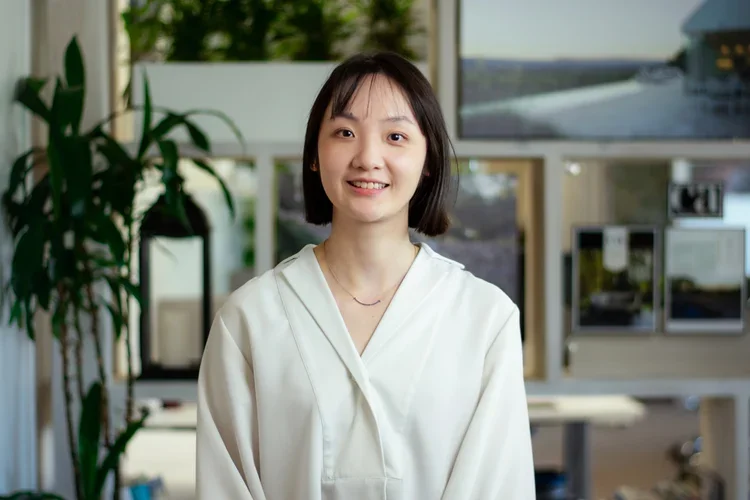 A young Asian woman with shoulder-length black hair, wearing a white blouse, standing in an office with creative artworks and a potted plant in the background.