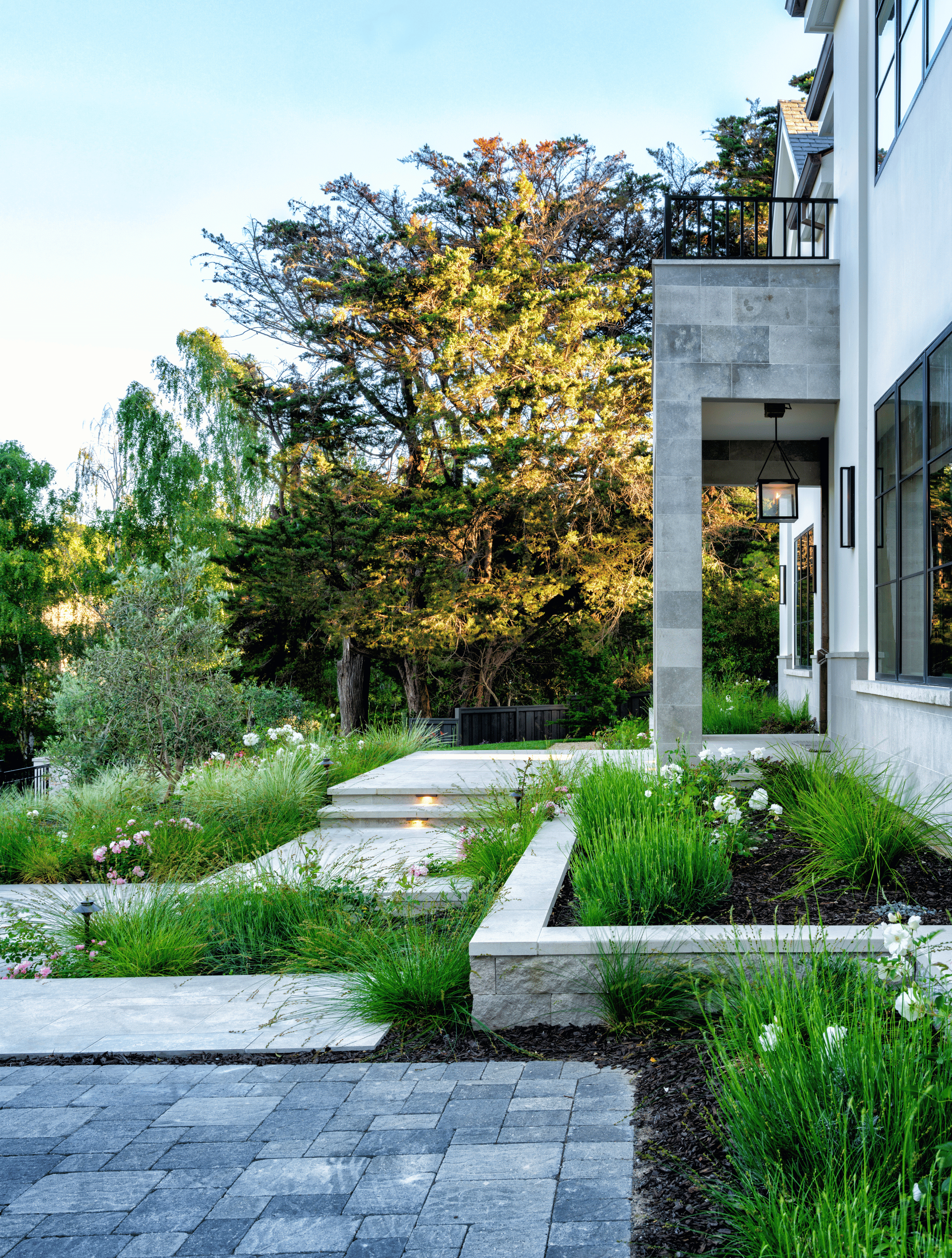 Modern house with a landscaped front yard featuring stone steps, lush green plants, and mature trees in the background, with a light sky above.