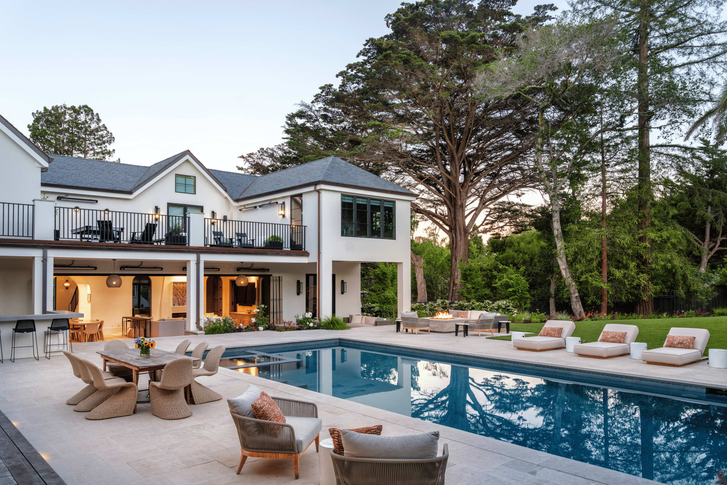 A modern two-story house with a backyard featuring a swimming pool, outdoor lounge area, dining table, fire pit, and lush green trees in the background