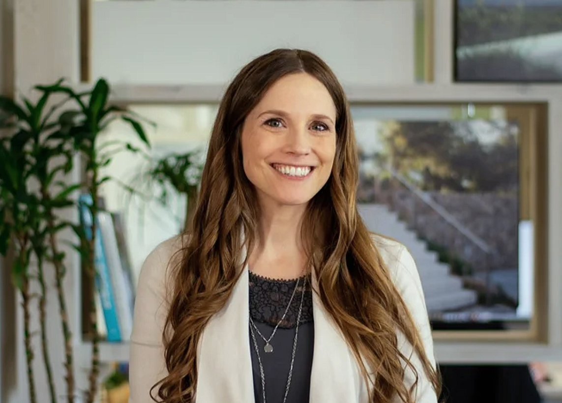 A woman with long, wavy brown hair smiling in an office environment, with plants and books in the background.