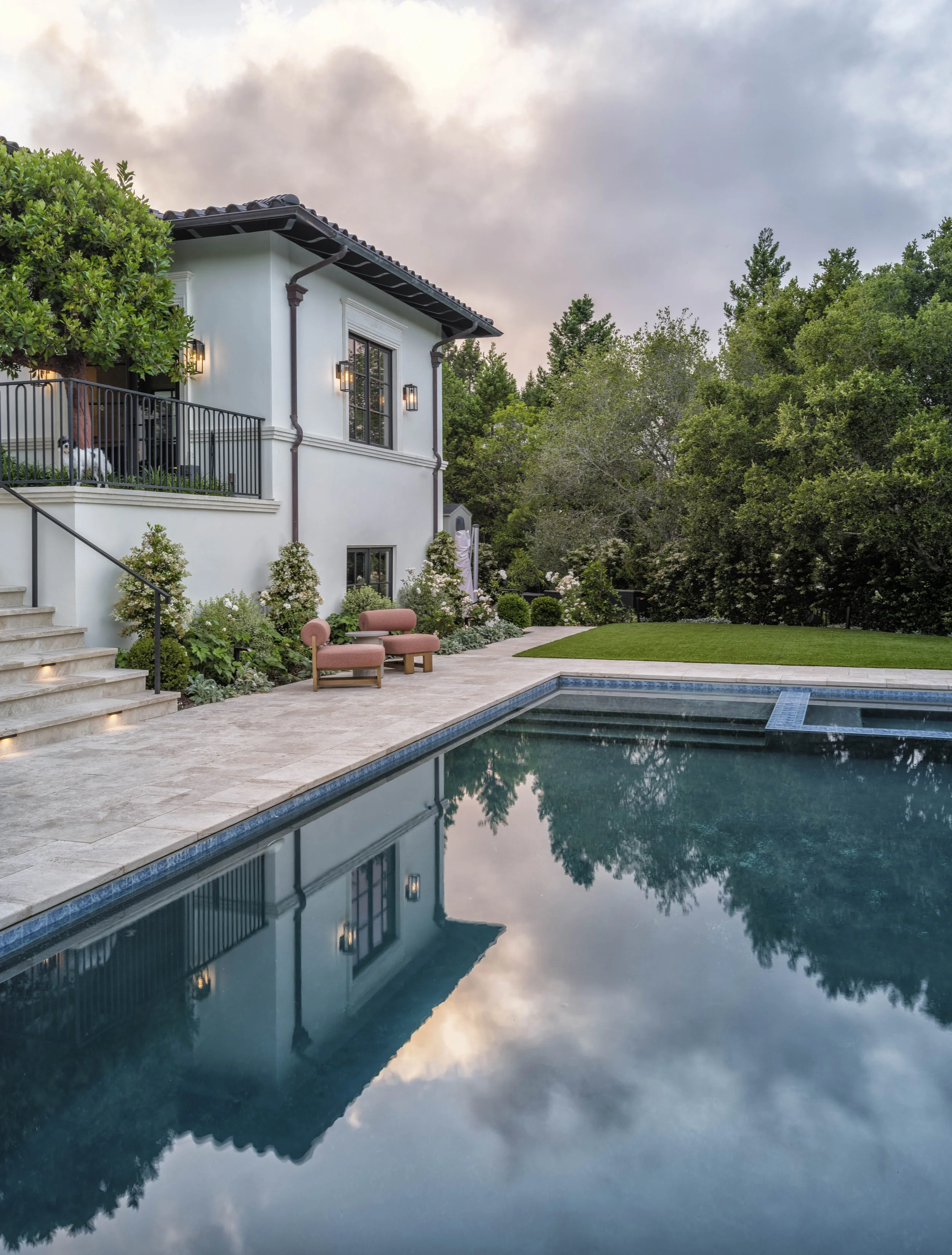 A backyard with a swimming pool, outdoor seating, lush greenery, and a white house with black trim and exterior lighting, under a partly cloudy sky.