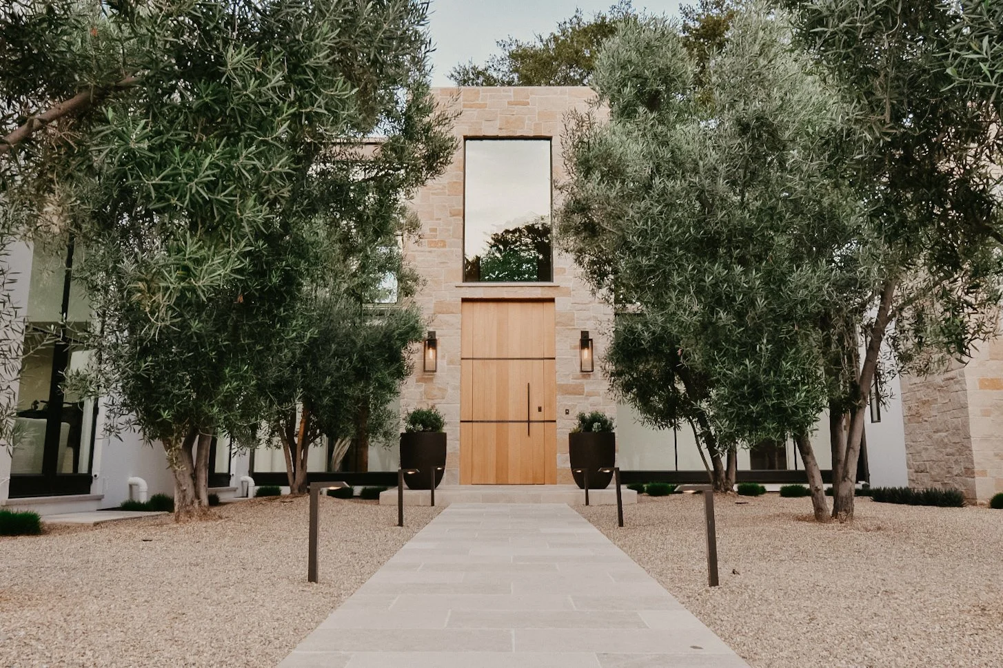 Modern house with stone exterior and wooden double door, flanked by two large potted plants. A stone pathway leads to the entrance, surrounded by gravel and trees. A large window reflects the sky and trees.