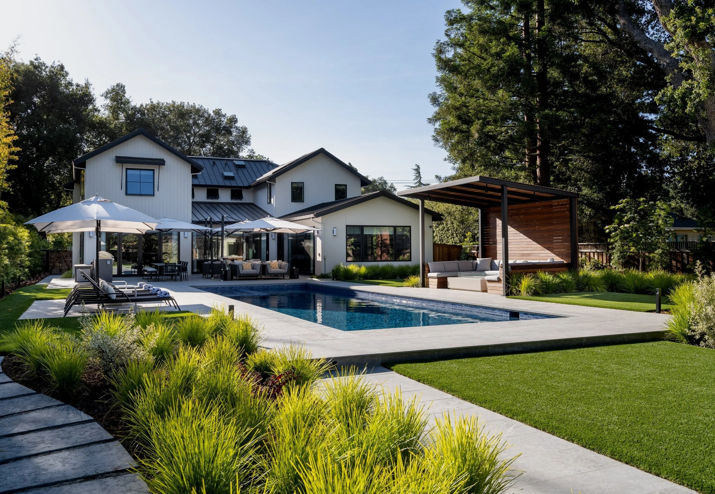 Modern backyard with a rectangular swimming pool, outdoor lounge area with umbrellas, covered seating area, and a well-maintained garden with green grass and shrubs.