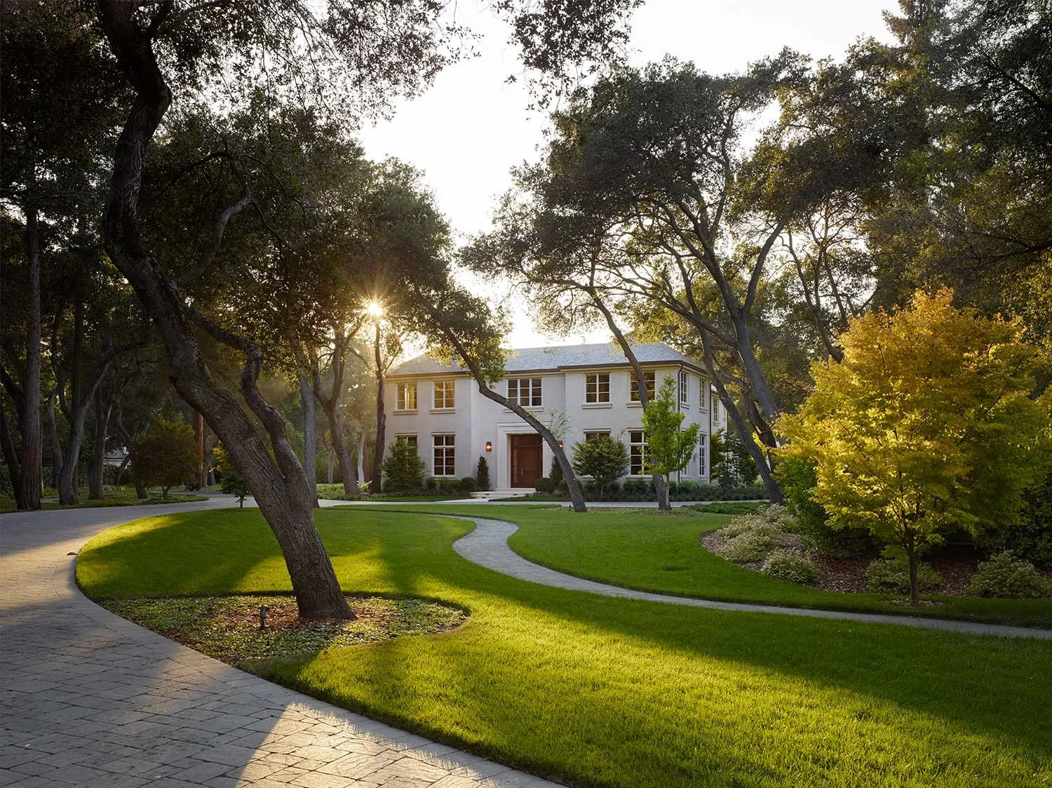 A white two-story house with many windows, surrounded by a lush green lawn, trees, and a winding stone pathway, with the sunlight shining through the trees.
