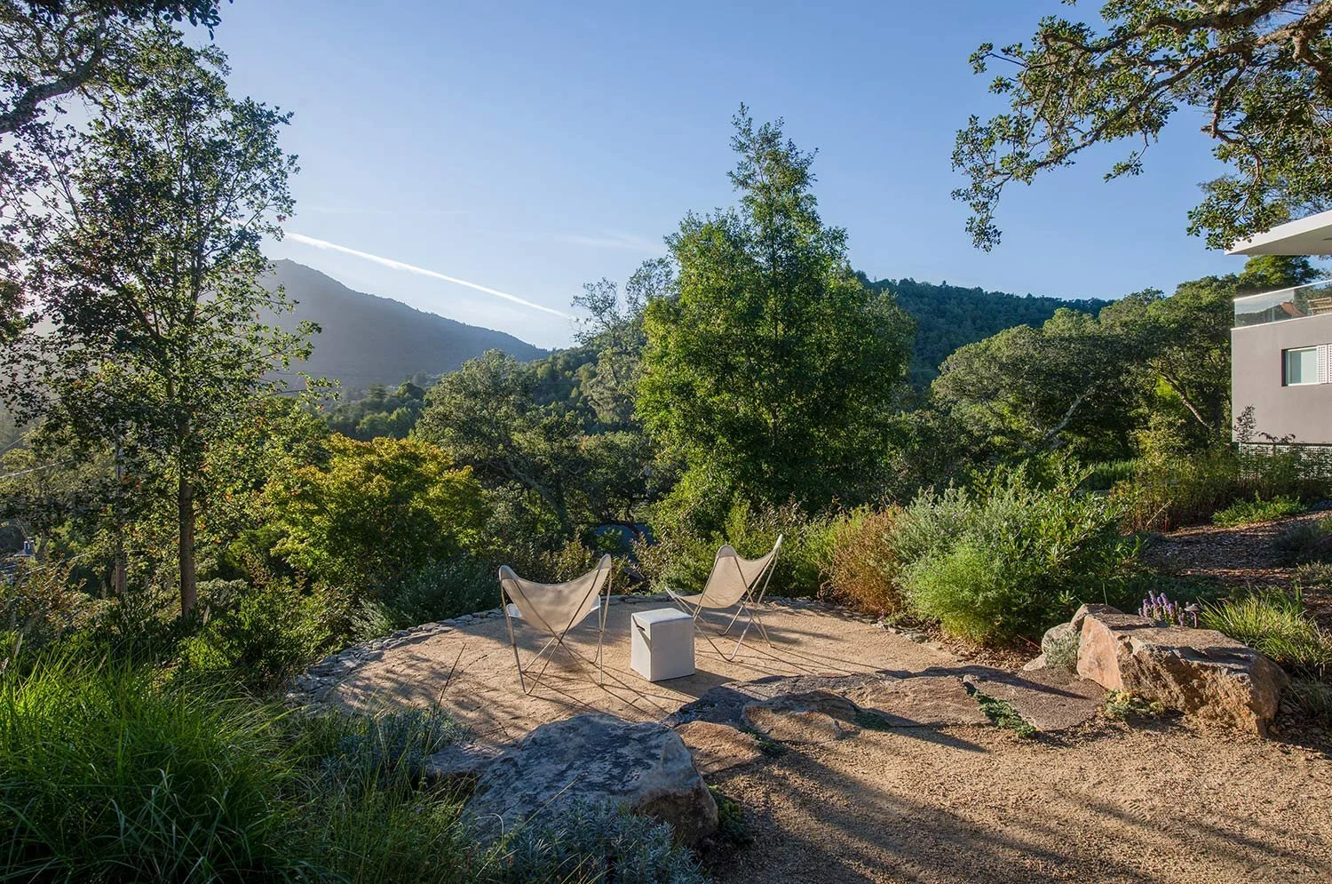 Two modern chairs with a small white table in a garden on a rocky area, surrounded by trees and plants, with mountains in the background under a clear blue sky.