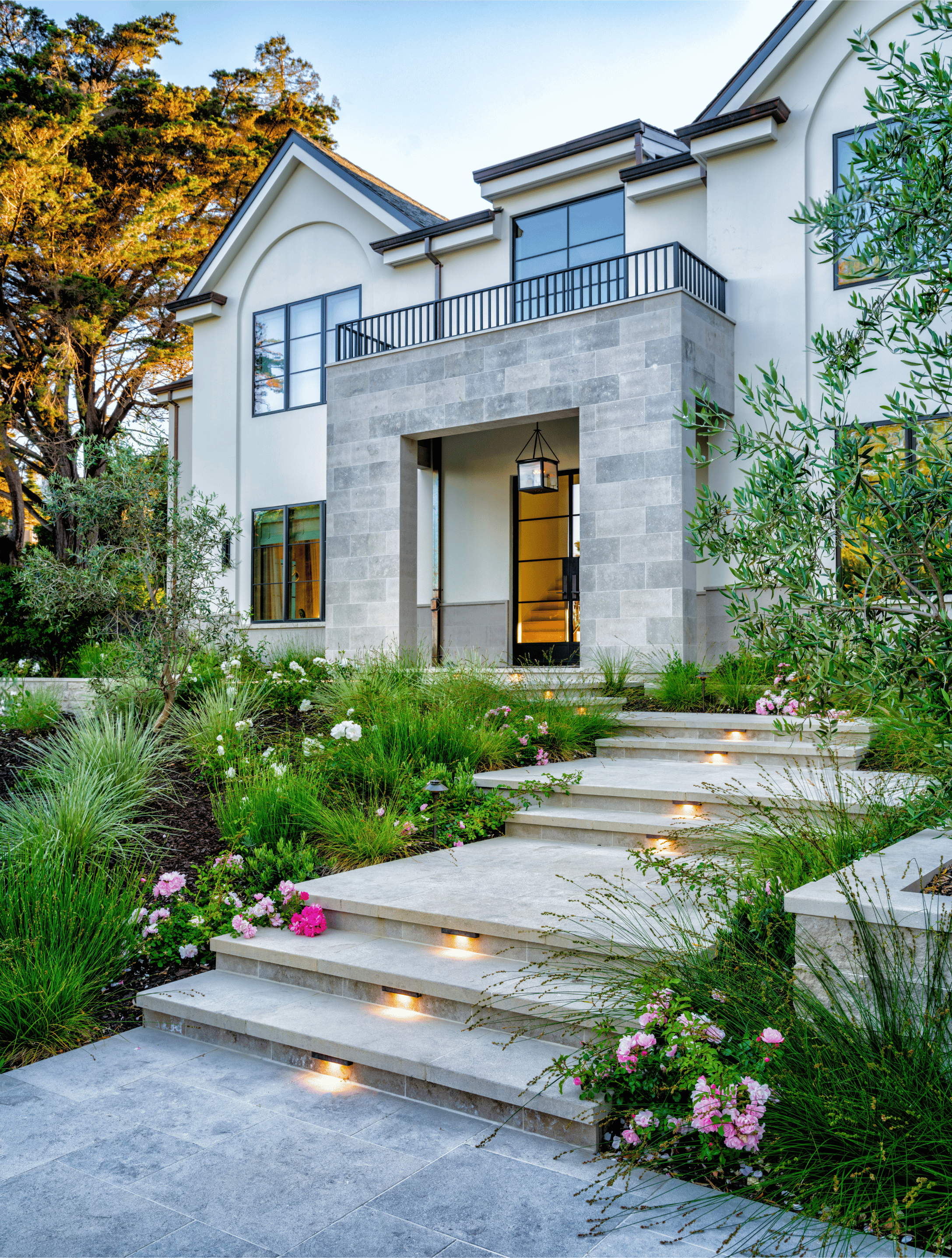 Modern white house with a front porch, exterior lighting, and landscaped garden with flowers and shrubs.