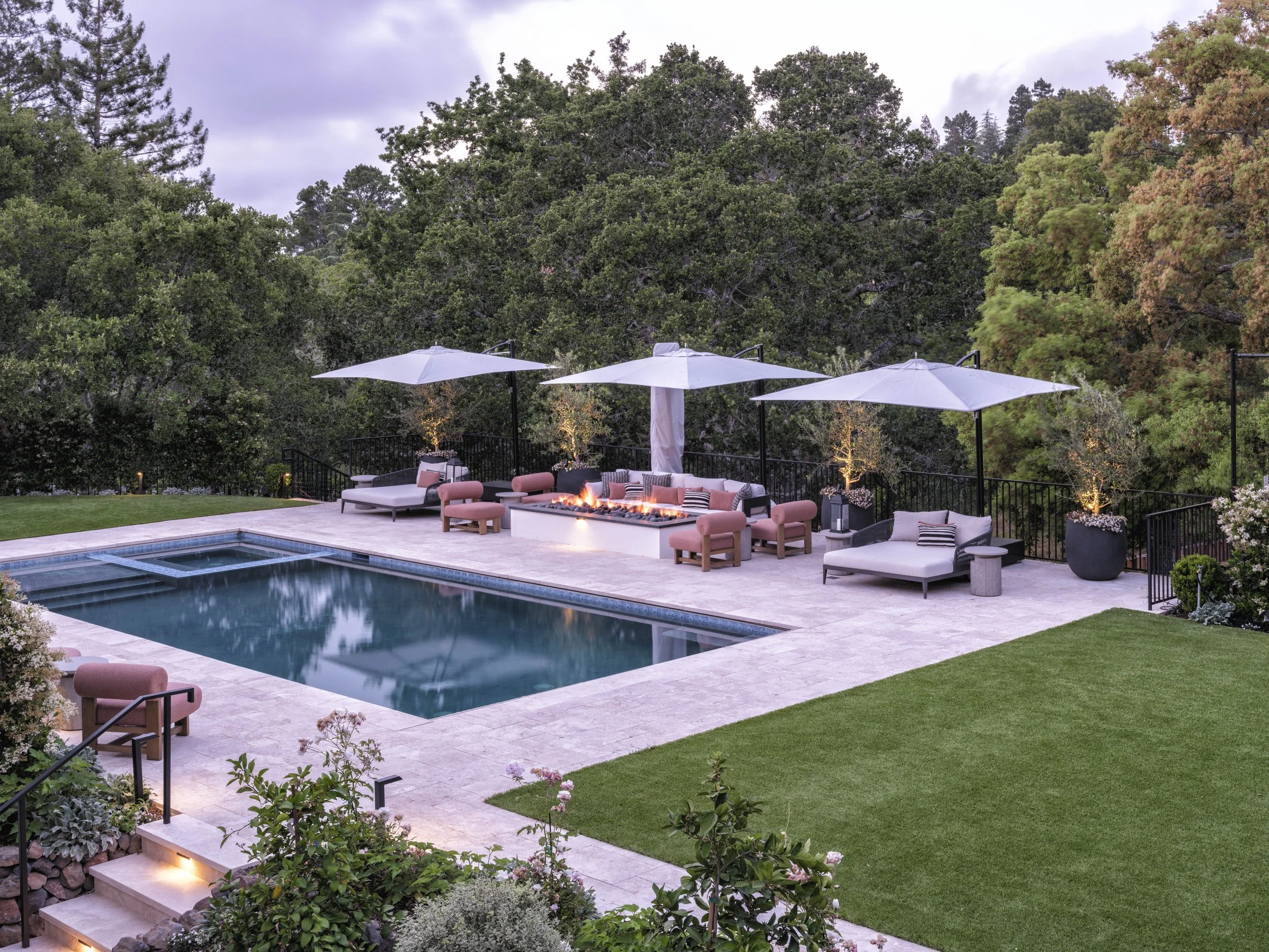 A backyard with a swimming pool, outdoor seating, umbrellas, and a fire feature surrounded by greenery and trees.